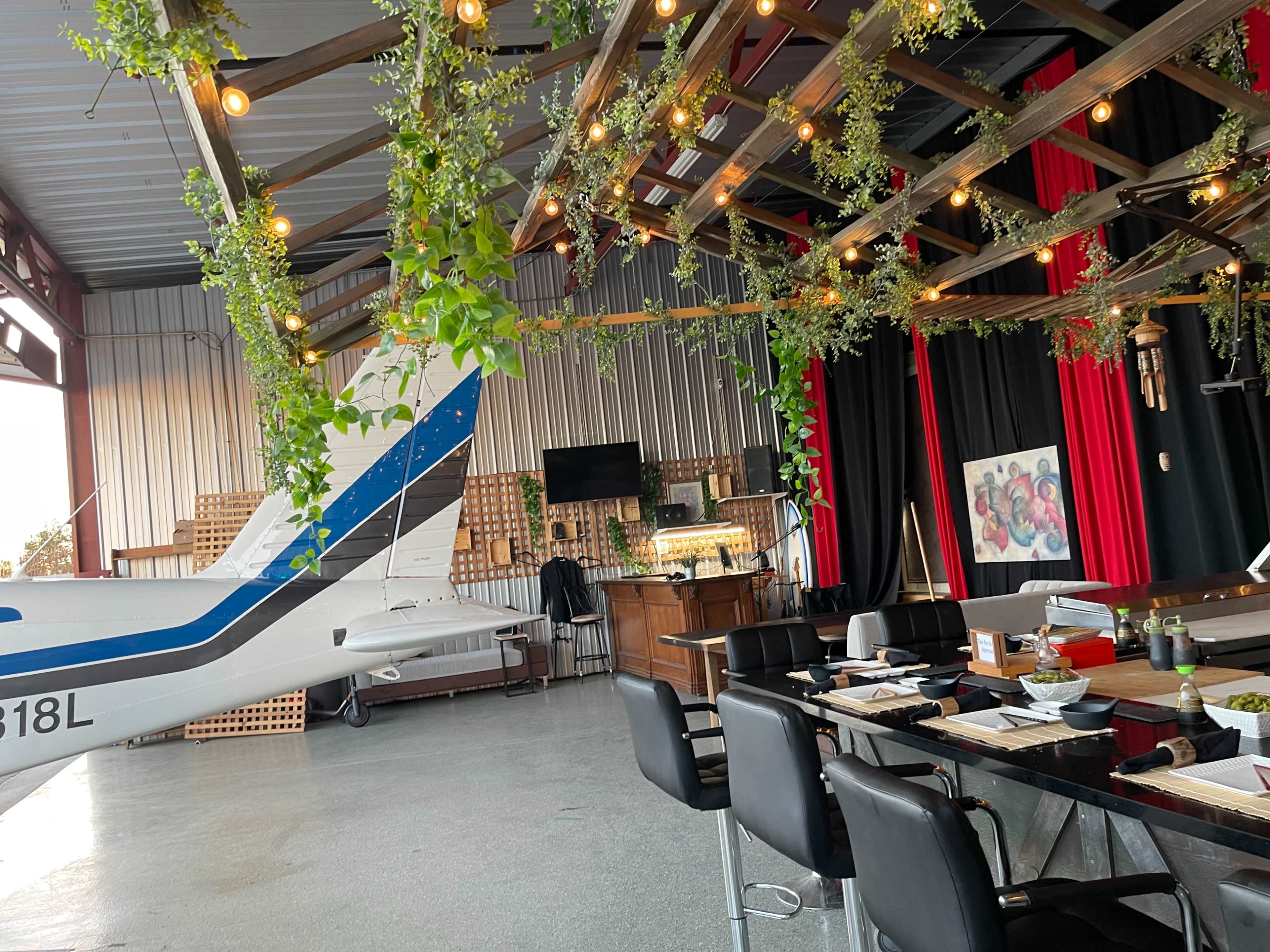 The interior features a modern dining area with a small aircraft parked nearby, decorated with hanging plants and string lights.