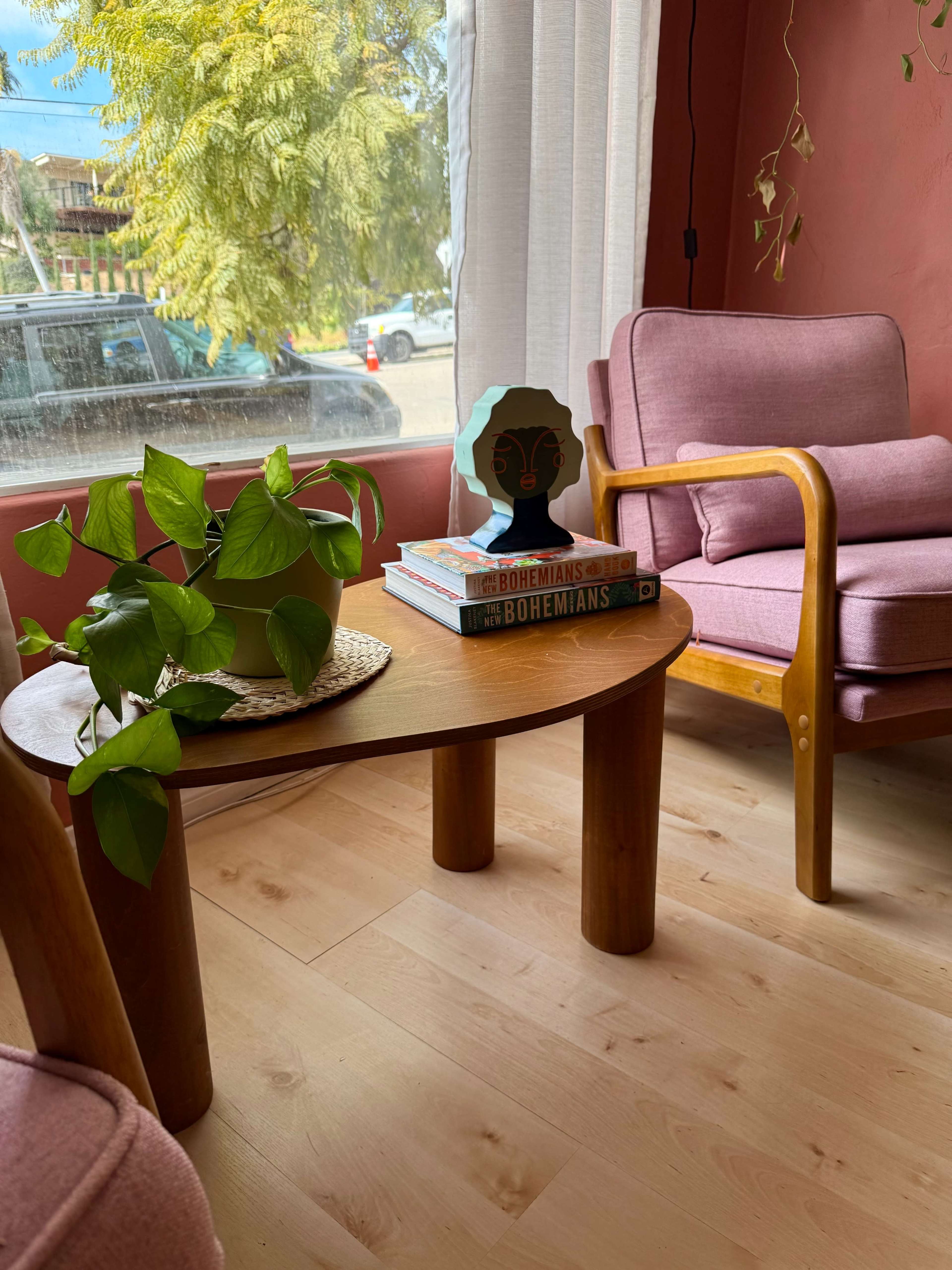 A small wooden table with a potted plant and books is positioned next to a pink armchair in a room with large windows and a view of the outdoors.