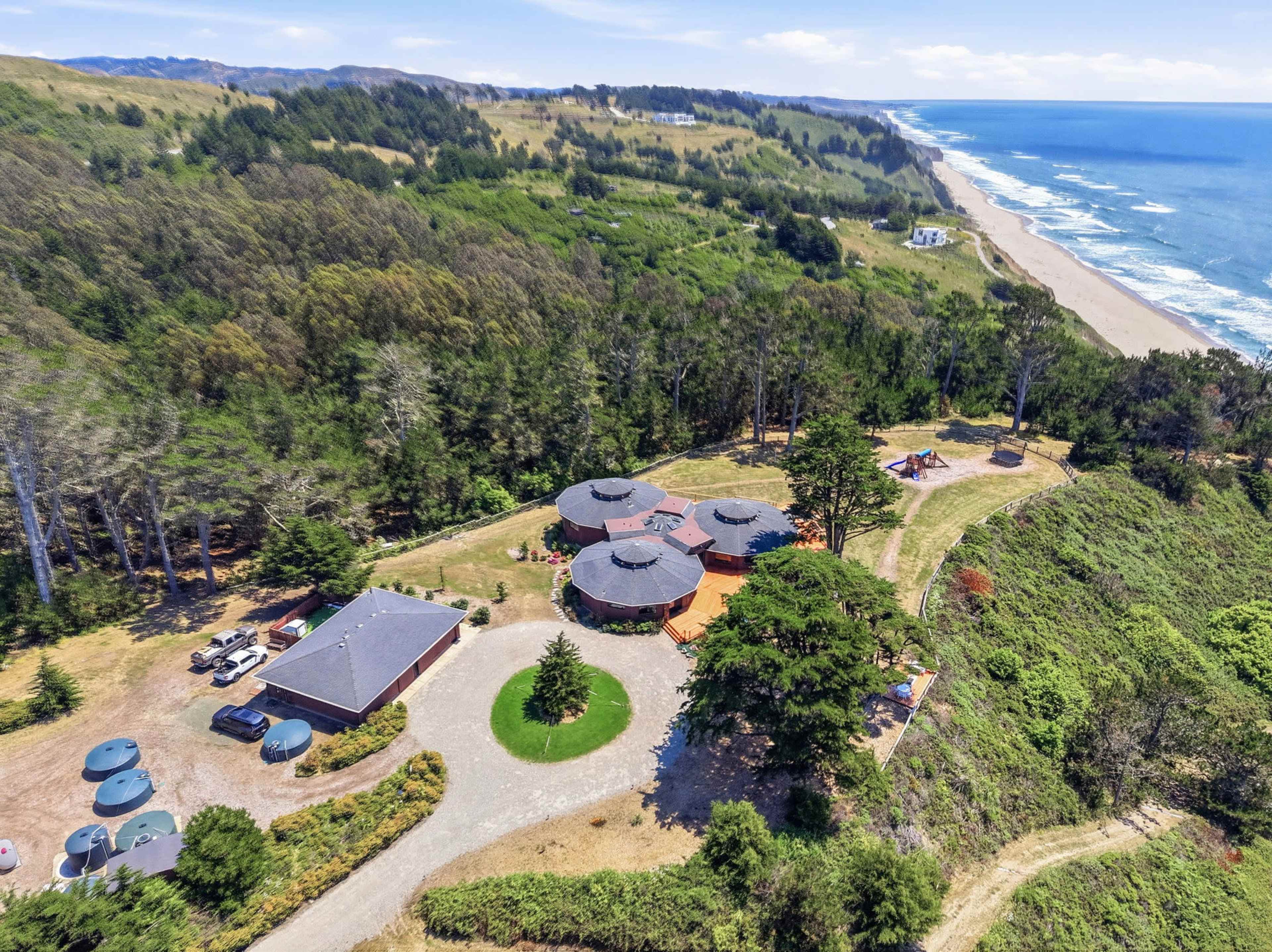The image shows an aerial view of a coastal property featuring circular buildings, surrounded by trees and terrain, with a beach and ocean visible in the background.