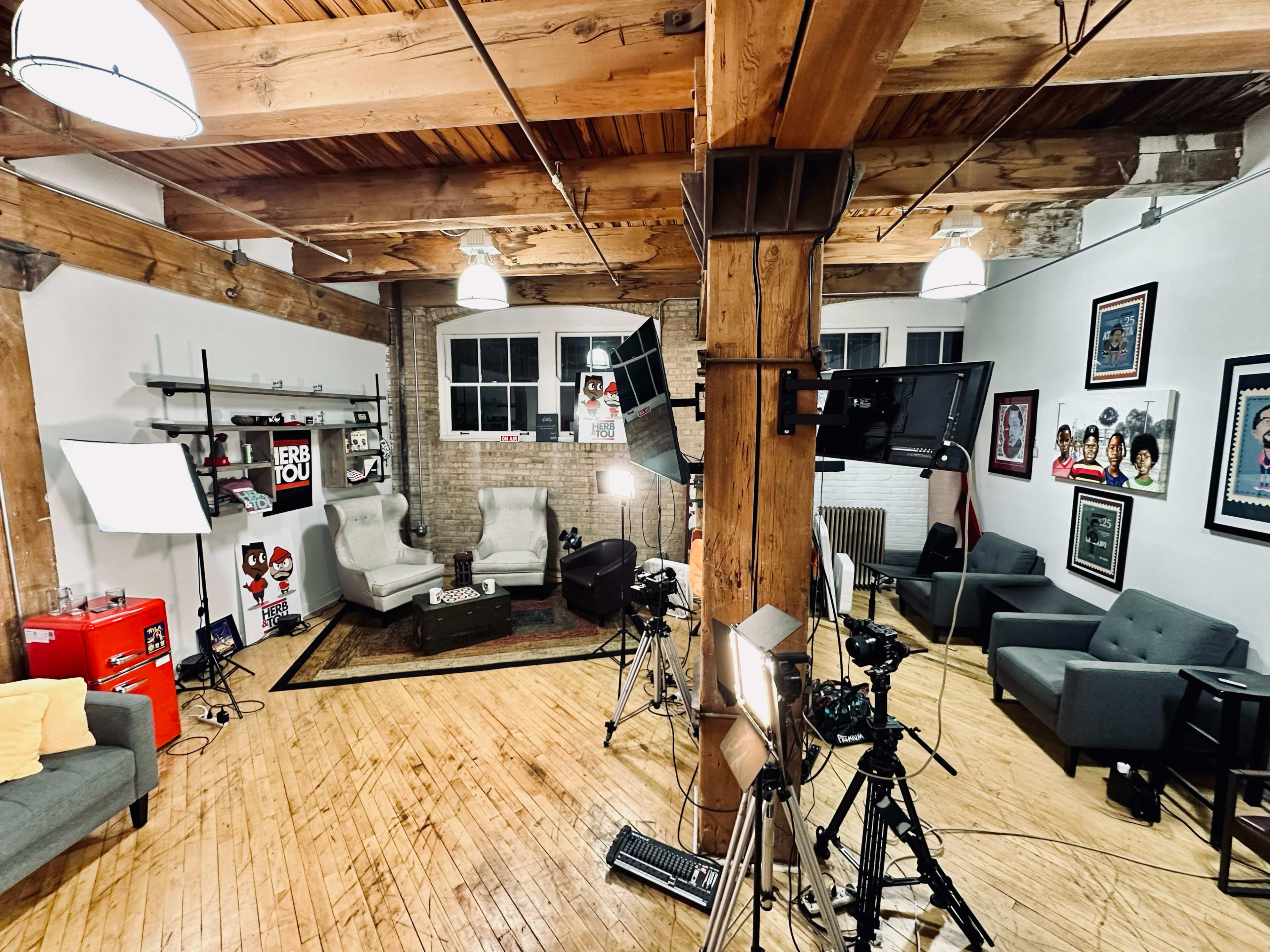 The image shows a spacious studio with wooden beams and a combination of comfortable seating areas, lighting equipment, and cameras set up for filming.