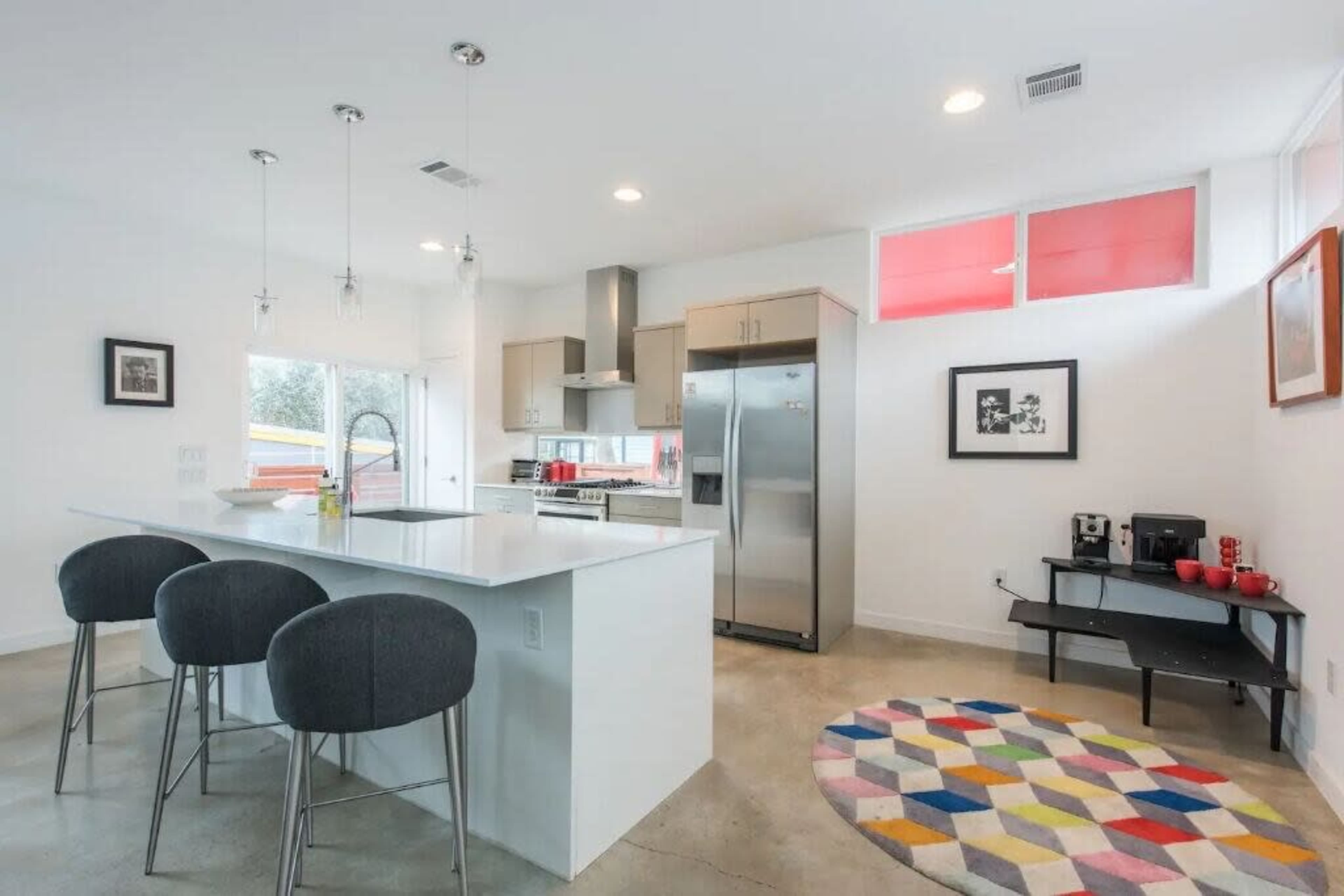 A modern kitchen features an island with bar stools, stainless steel appliances, and colorful decor including a patterned rug.