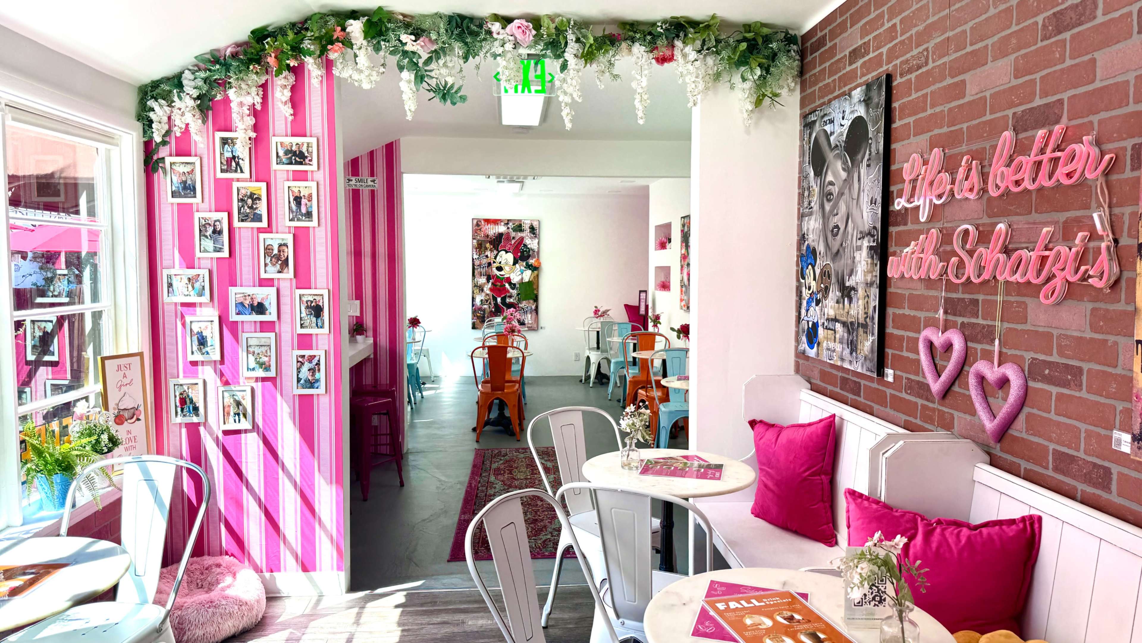 The image shows a brightly decorated café with pink and white walls, floral decorations, and wooden furniture arranged around small tables.