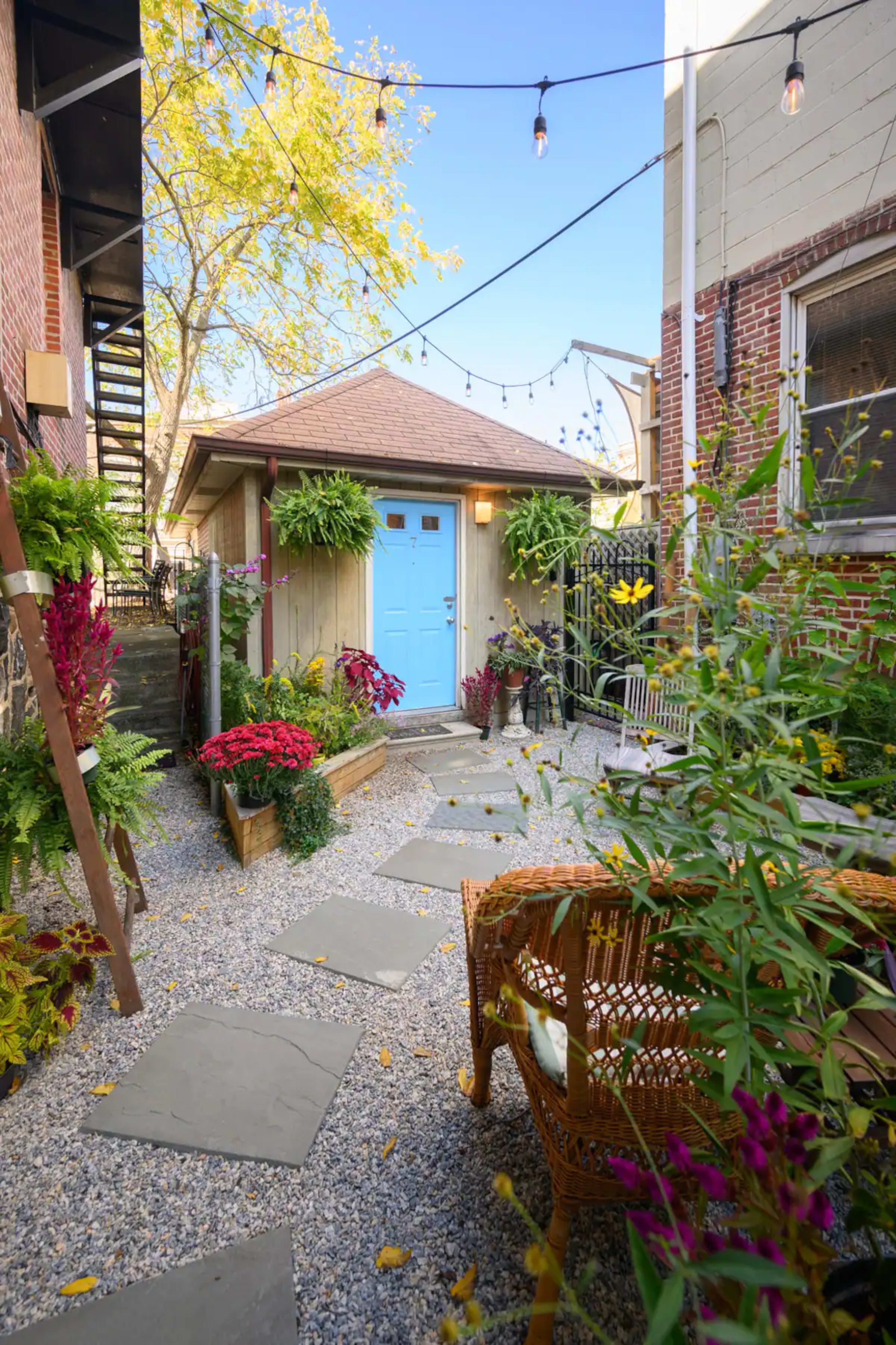 A small, gravel courtyard features a blue door, surrounded by planters and colorful flowers, with decorative string lights overhead.