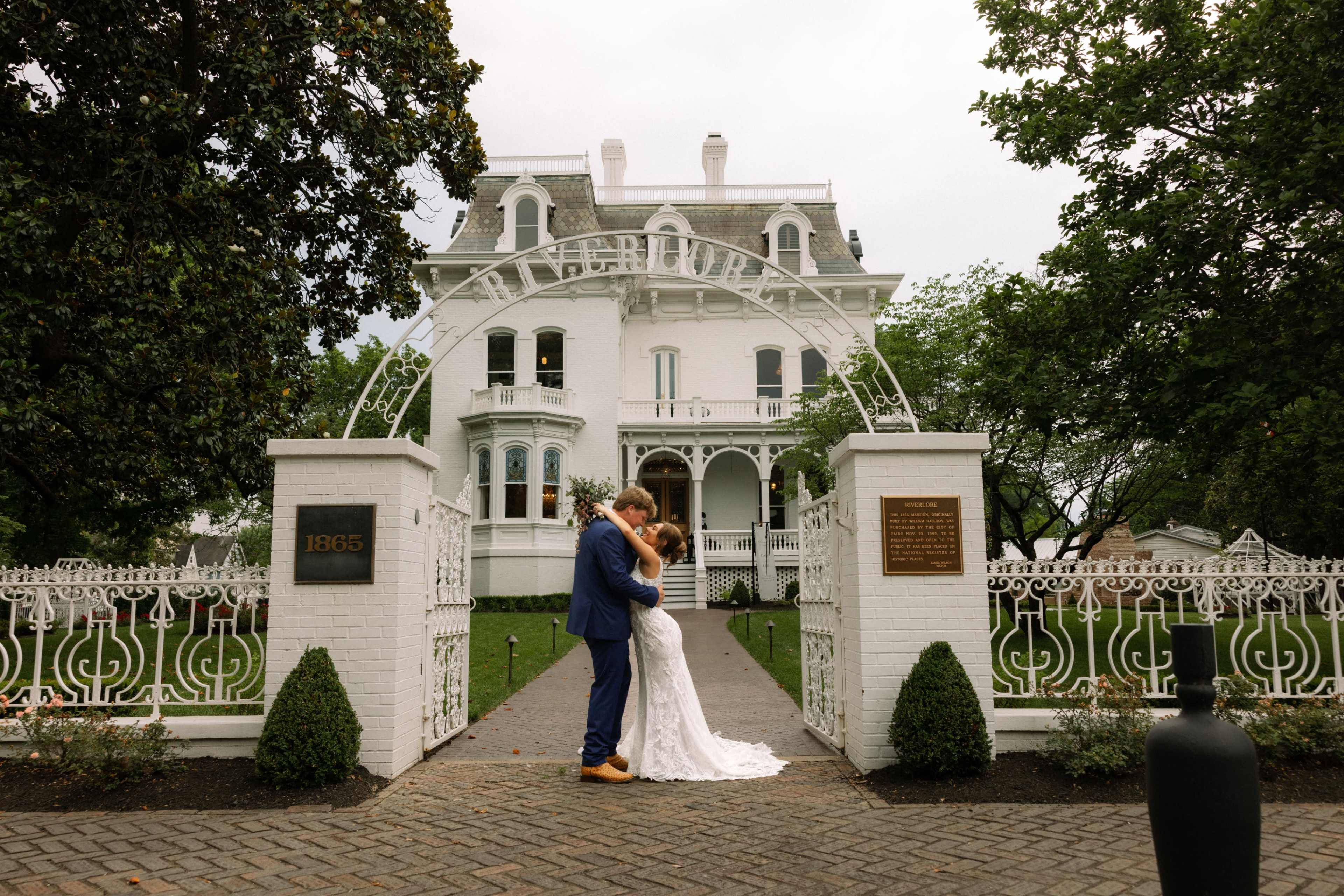 A newlywed couple embraces in front of a large white Victorian house with an ornate archway and grassy lawn.