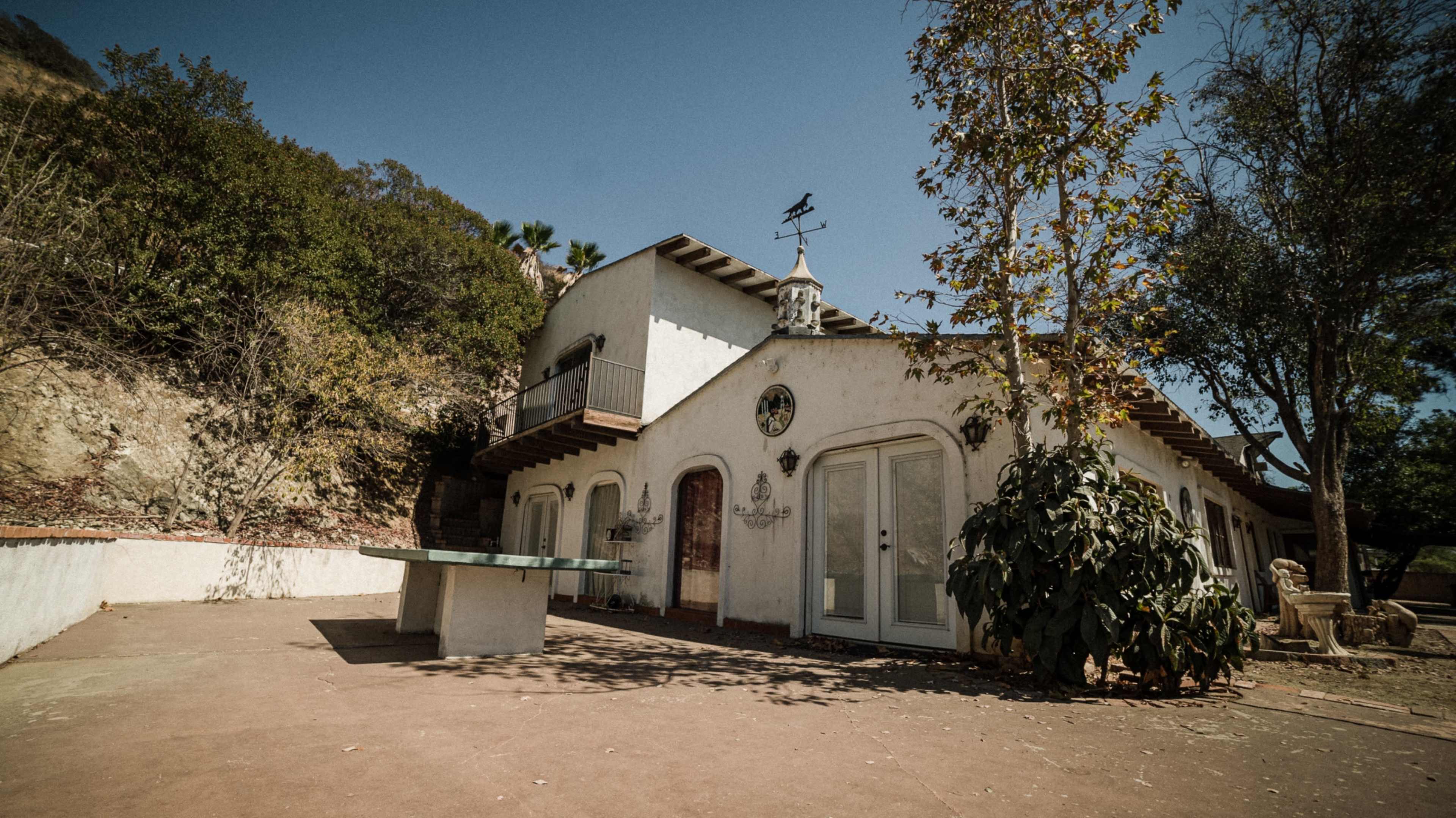 A stucco house with a tiled roof and a patio, surrounded by trees and hillside.