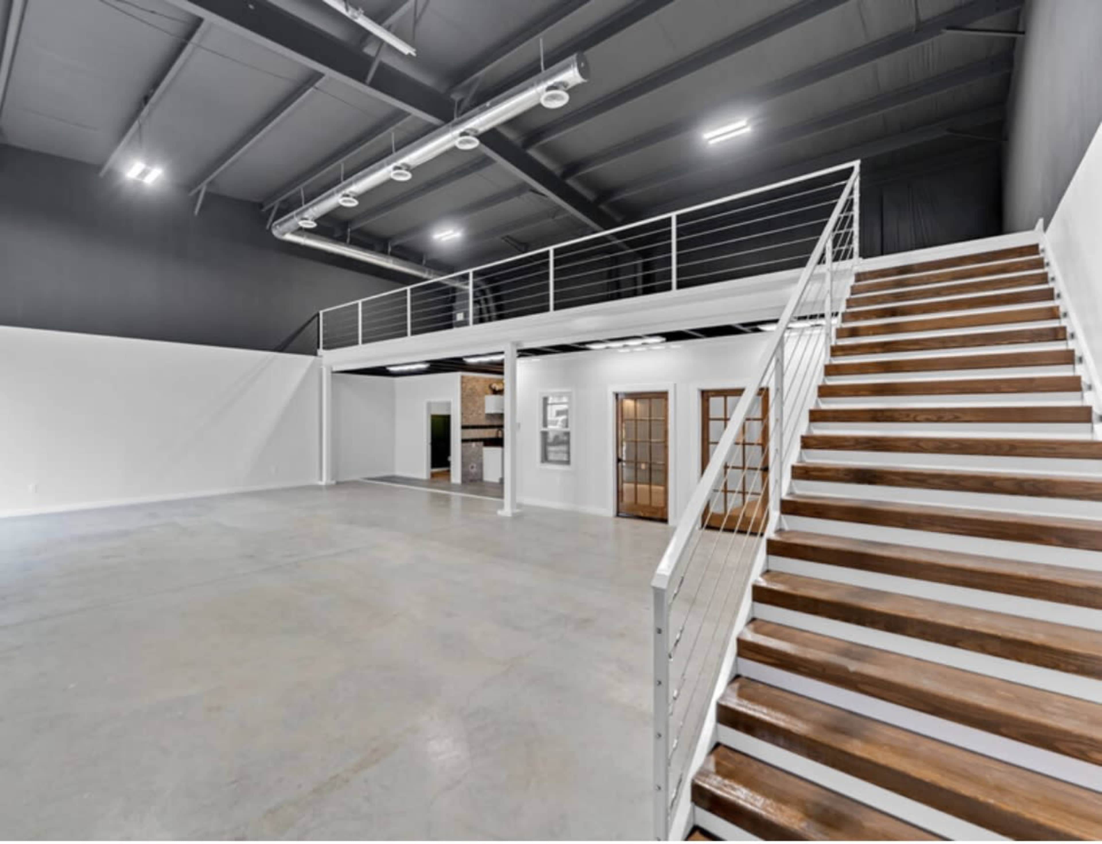 The image shows a spacious, modern interior with an upper loft and a staircase leading to it, featuring concrete flooring and large walls.