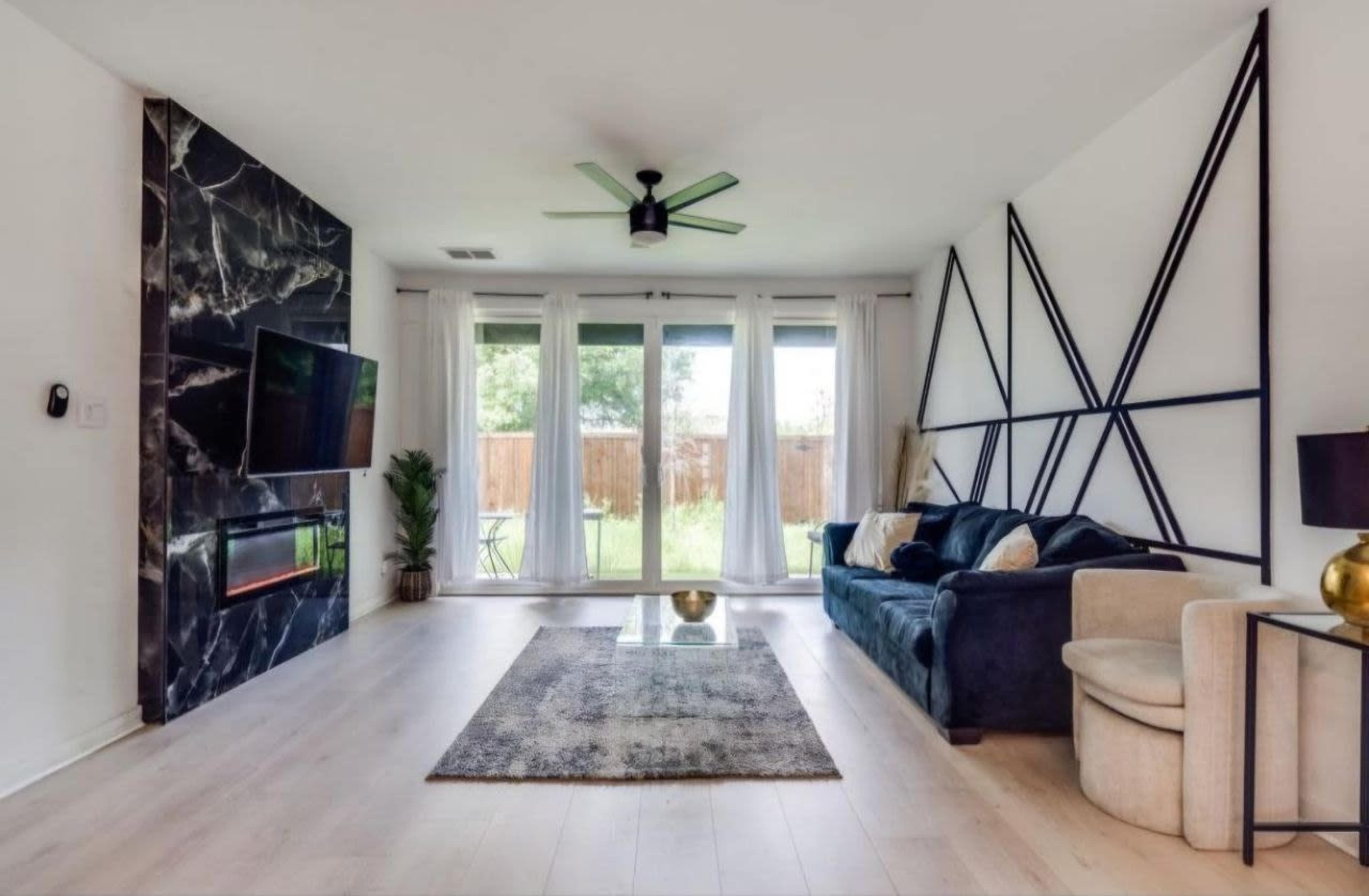 A modern living room featuring a dark marble accent wall with a television, a blue sofa, and large windows overlooking a backyard.