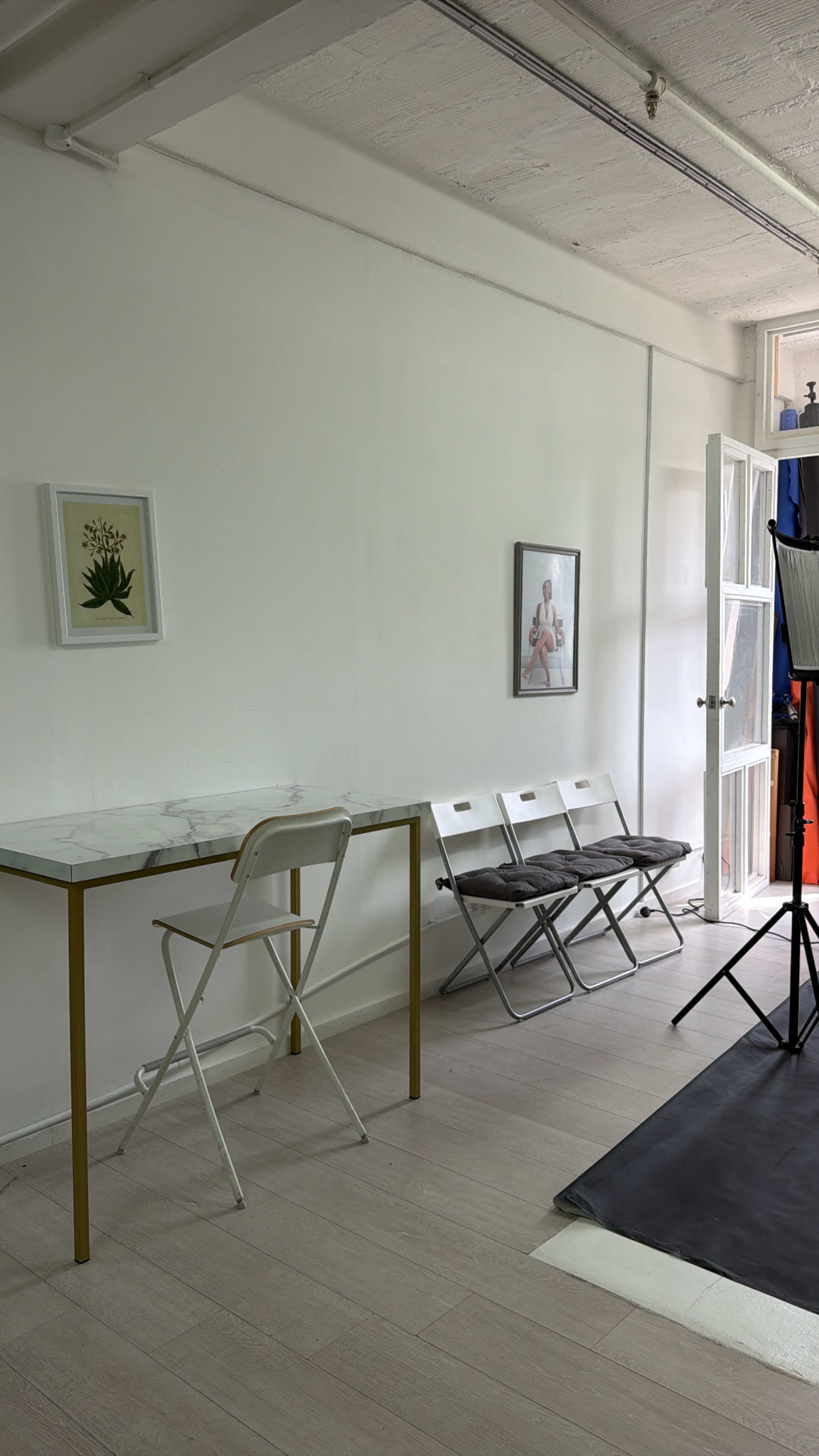 A small room features a marble-top table with a gold frame, three folding chairs against the wall, and a large window beside a photography light setup.