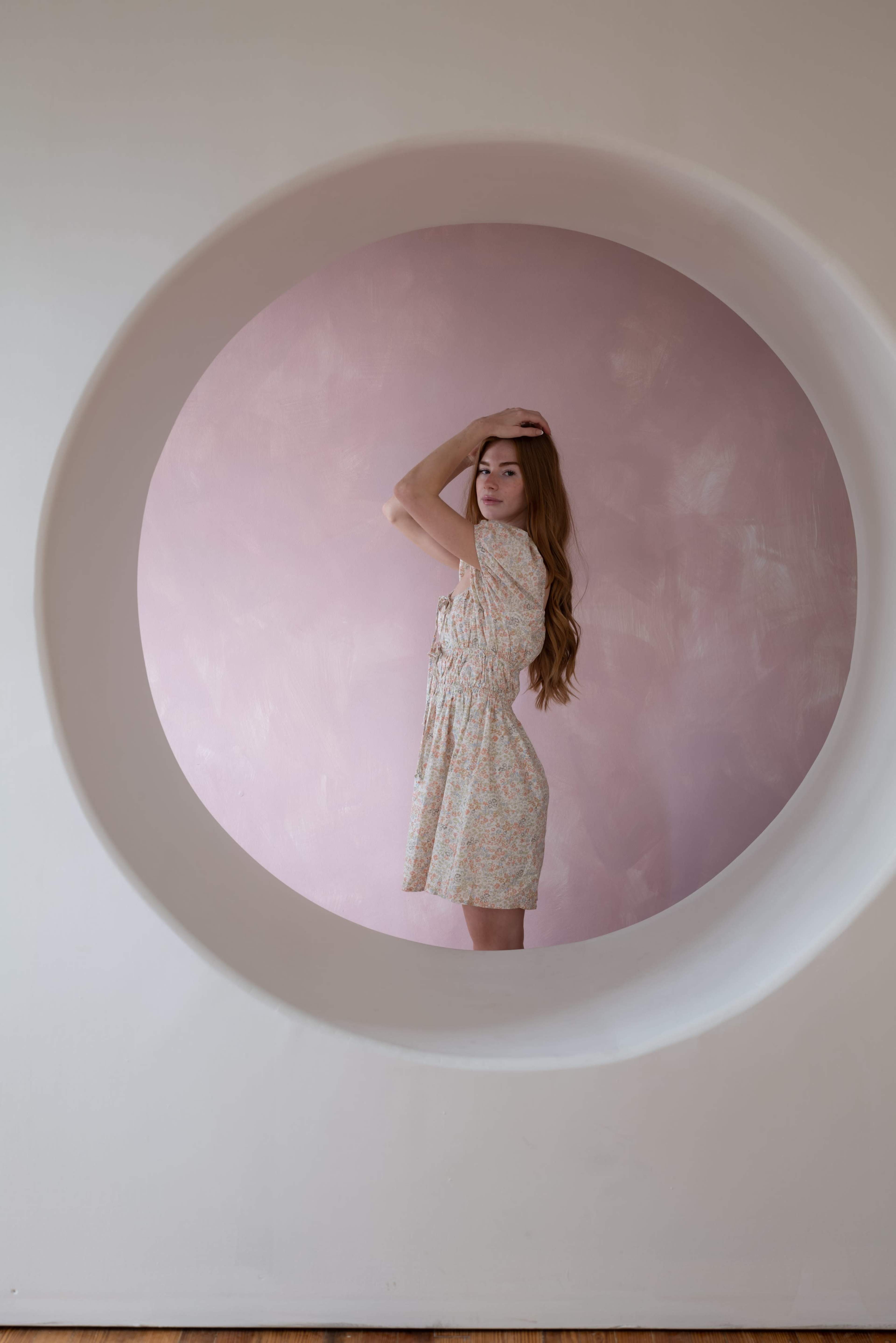 A woman with long hair poses inside a circular cutout in a wall, framed against a pink backdrop.
