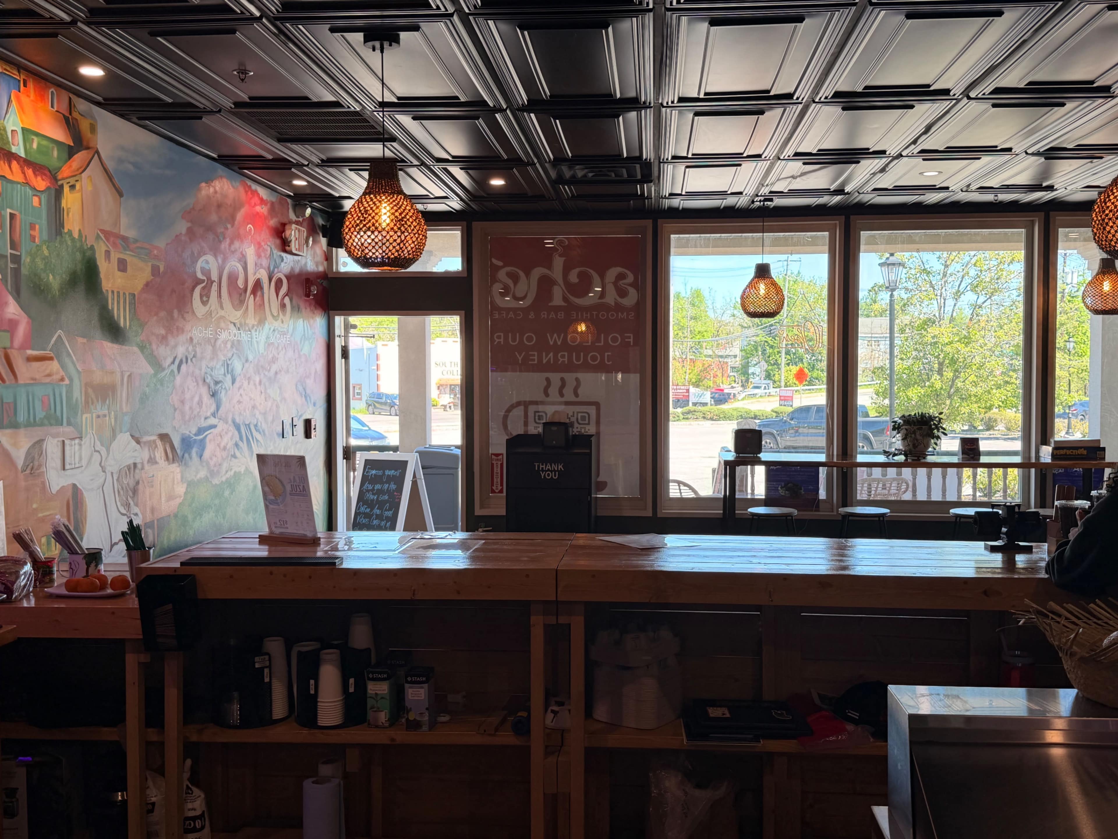 The image shows a cafe interior with wooden countertops, decorative light fixtures, and large windows that allow natural light to fill the space.