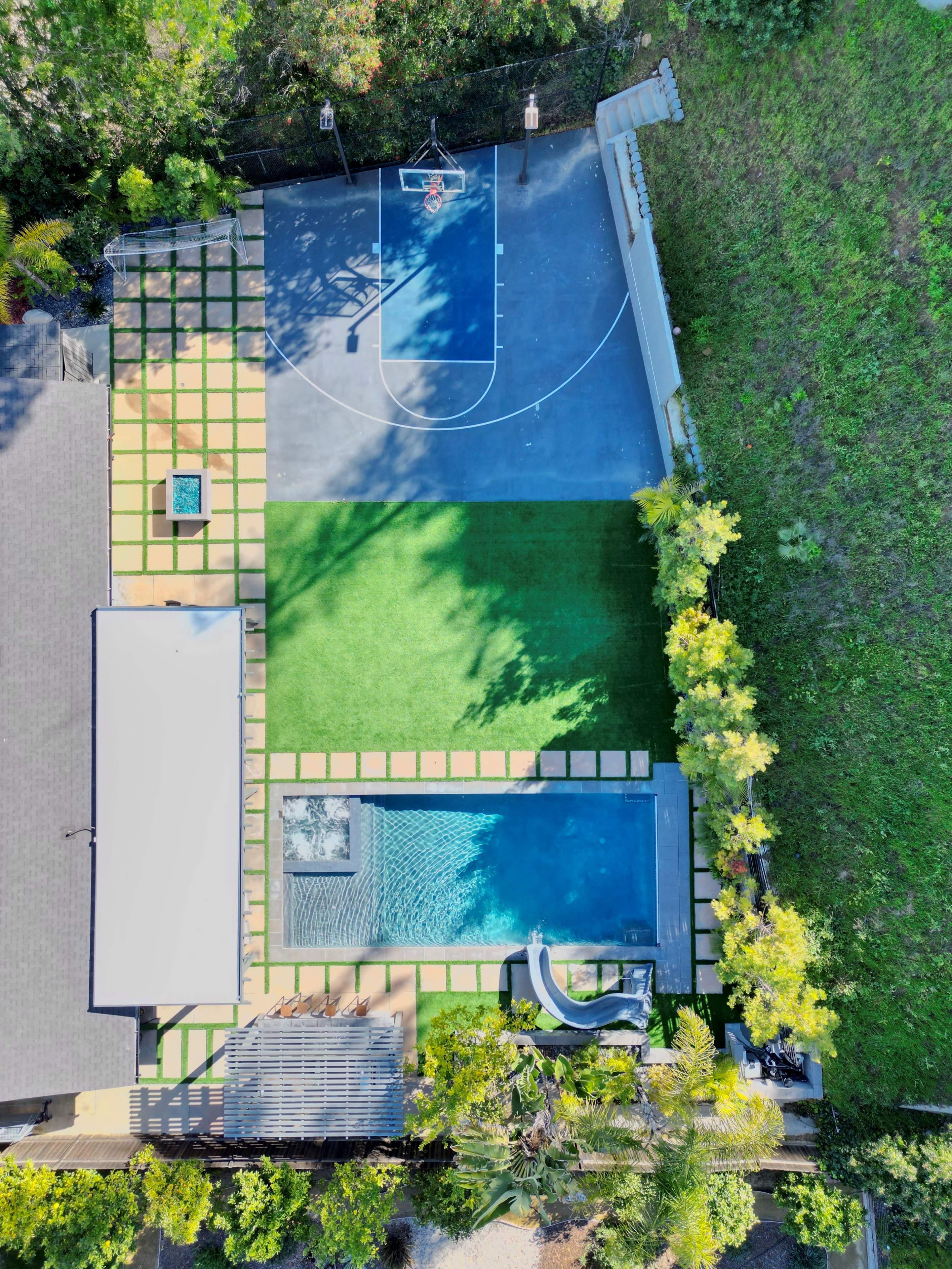 An aerial view of a backyard featuring a basketball court, a green lawn, and a swimming pool surrounded by landscaping.