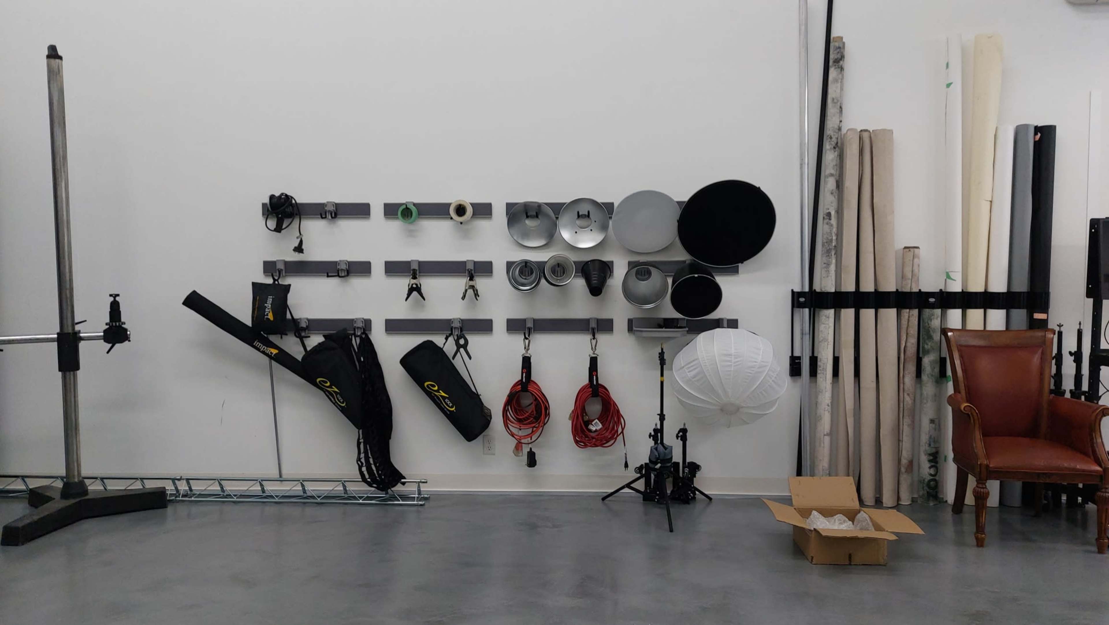 The image shows a wall-mounted storage arrangement with various photography equipment, including light modifiers, cables, and a white umbrella, alongside a cardboard box on the floor.