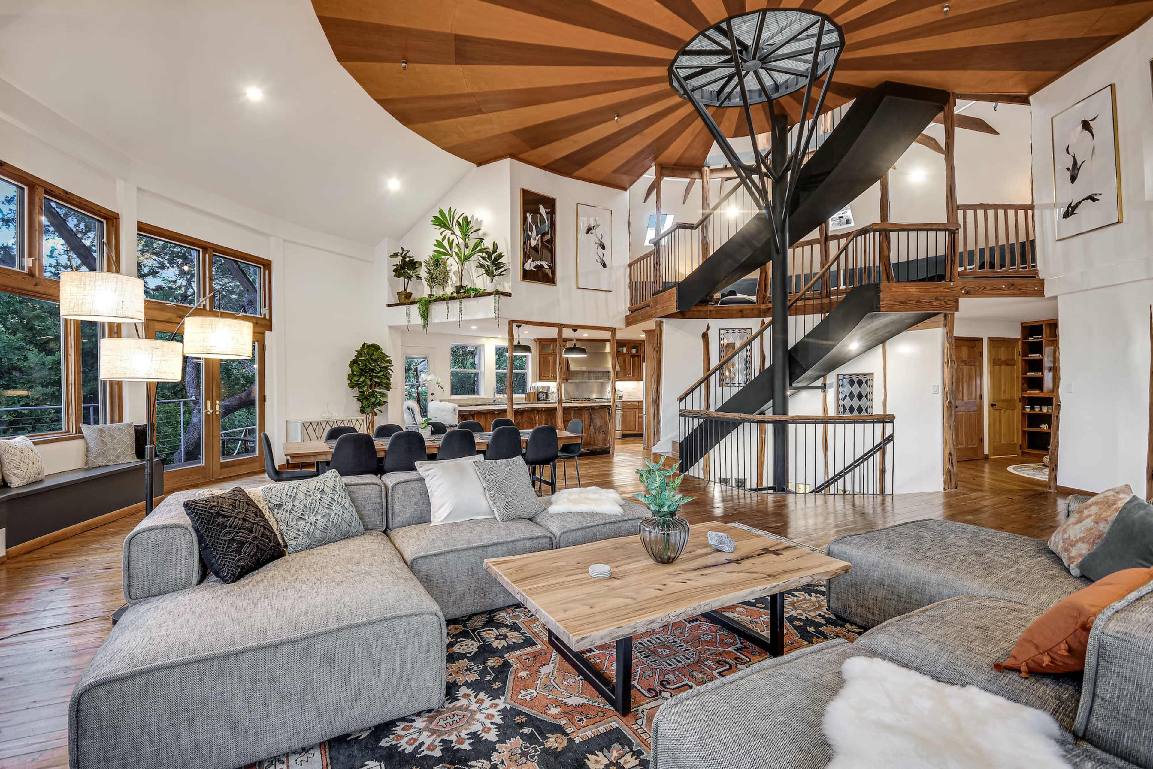 A spacious living area with a large sectional sofa, a wooden coffee table, and a spiral staircase leading to an upper level, surrounded by large windows.