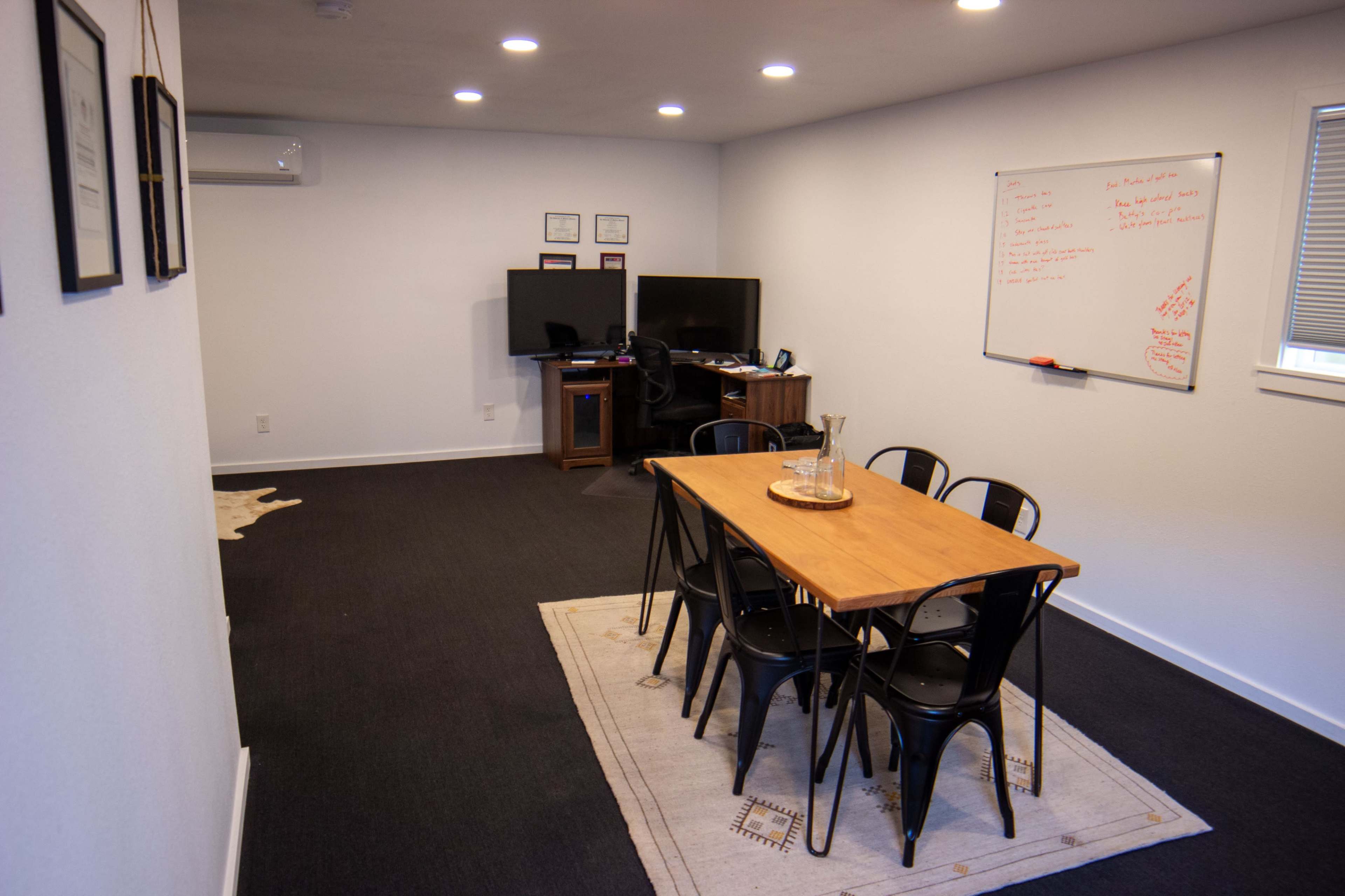 The image shows a light-colored room with a dining table surrounded by six black chairs, a whiteboard on the wall, and two computer monitors on a desk.