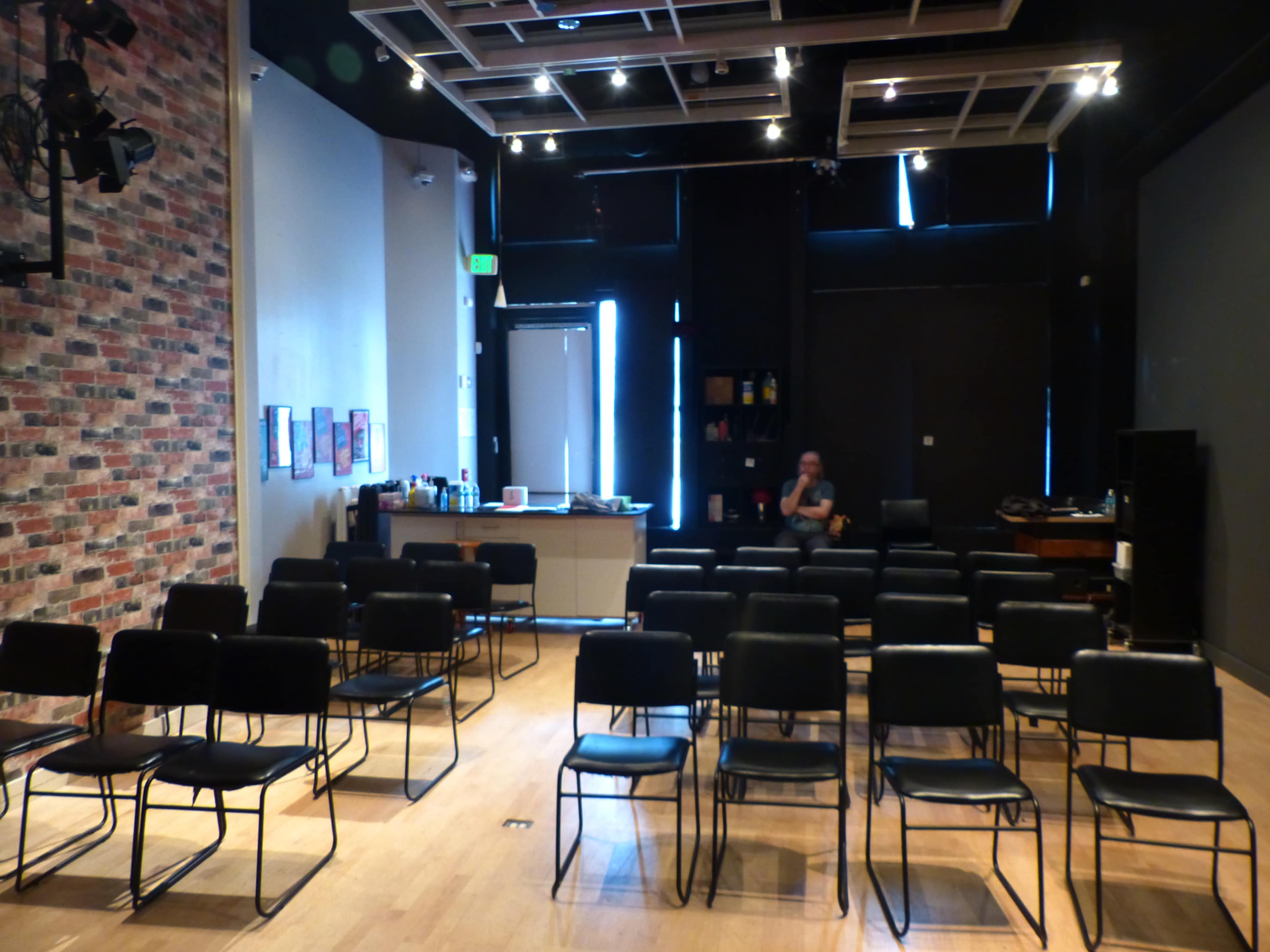 A nearly empty room with rows of chairs facing a stage, featuring a brick wall, and a lone person seated in the back.