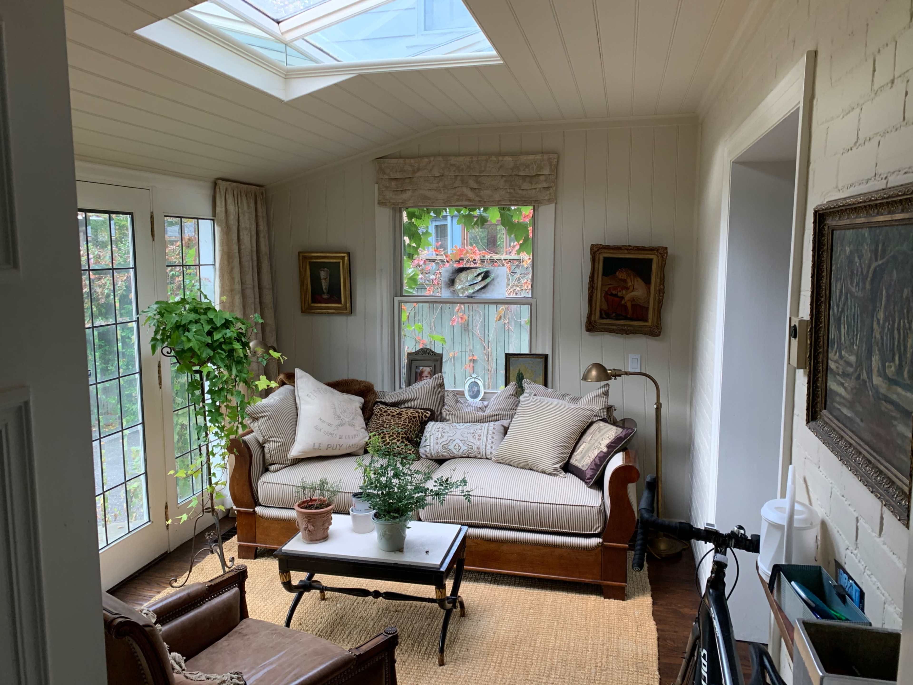 A sunlit corner of a room featuring a couch adorned with pillows, a small coffee table, and plants near large windows.