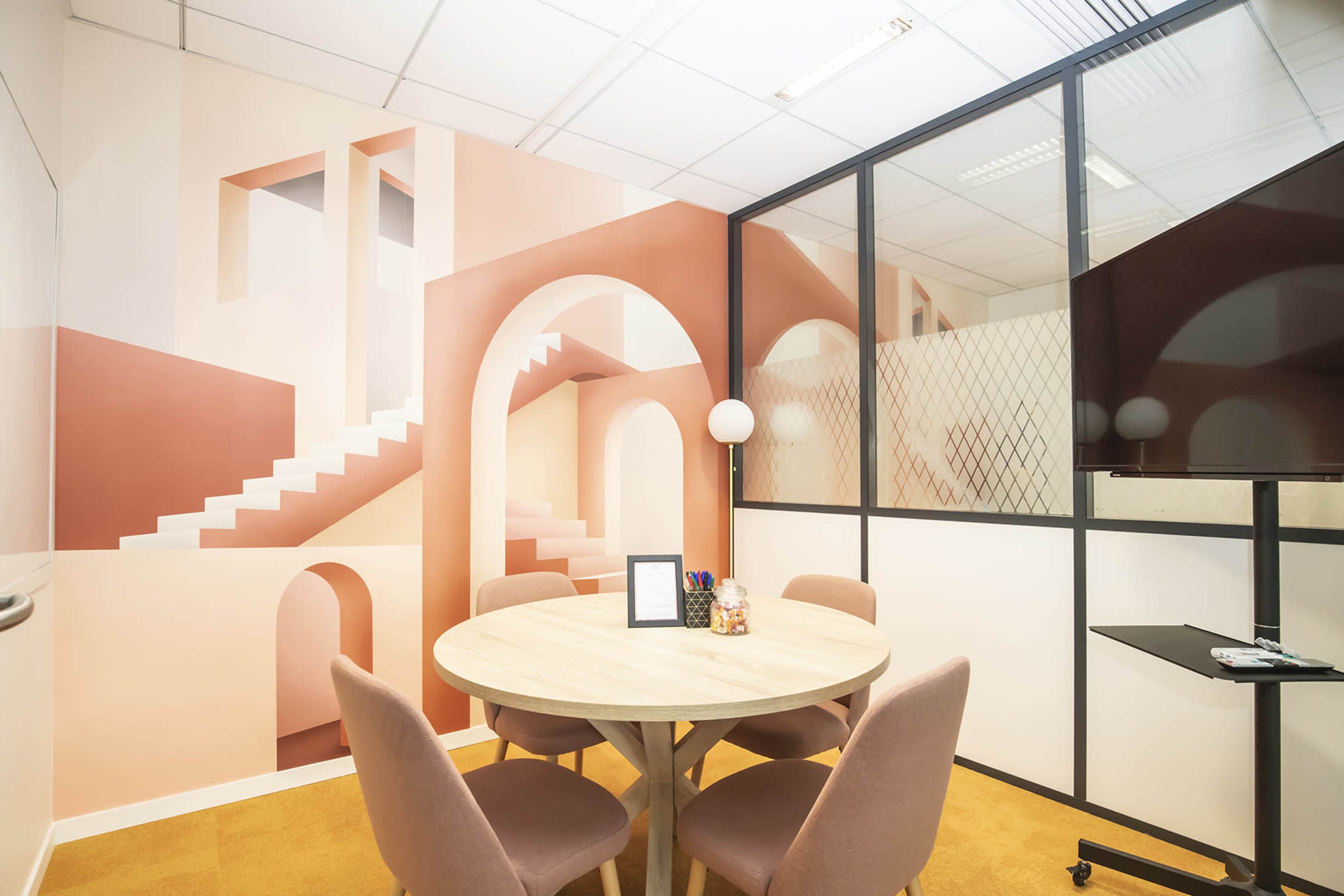 A modern meeting room features a round wooden table surrounded by upholstered chairs, with a mural of arches and stairs on the wall and a TV on a stand nearby.