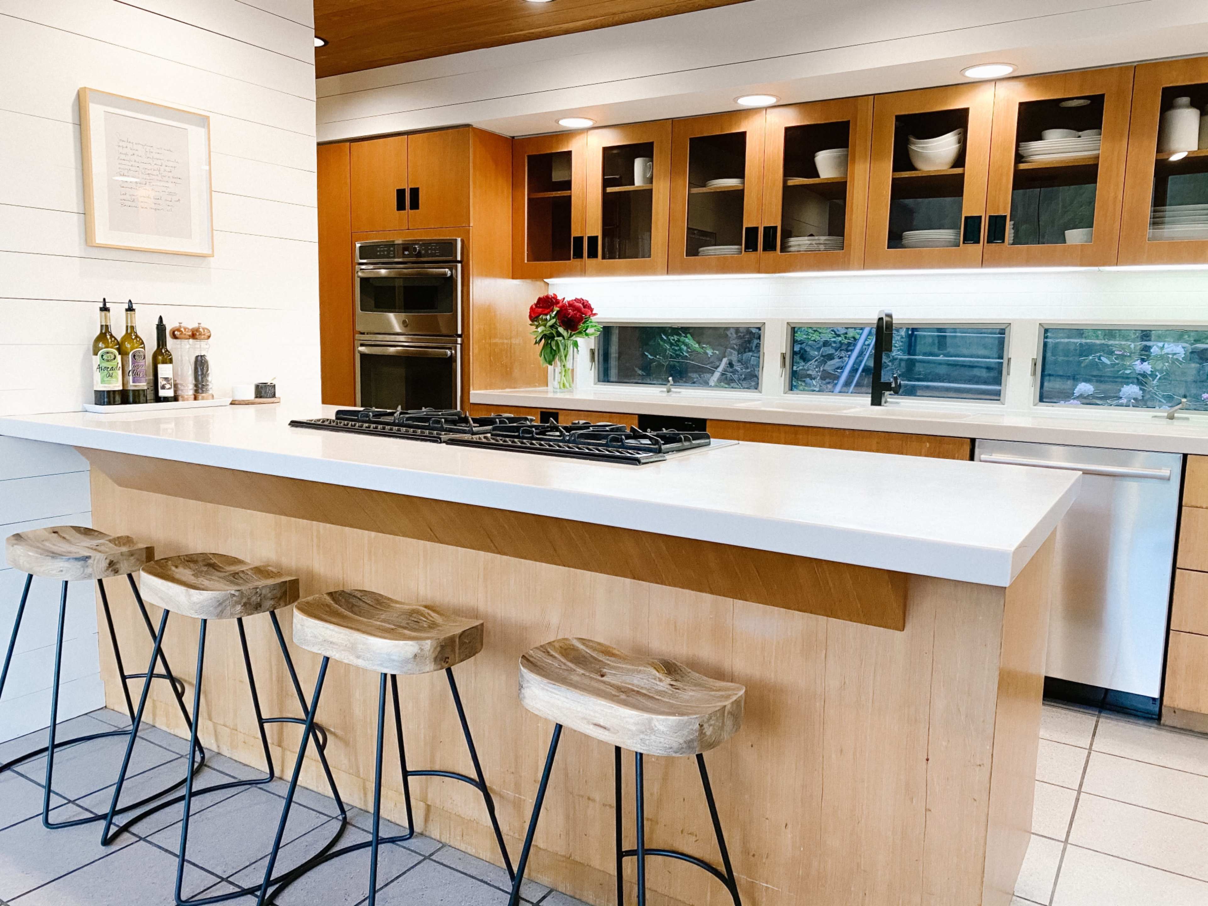 The kitchen features a central island topped with a white countertop and wooden stools, surrounded by wooden cabinetry and glass-fronted shelves filled with dishware.