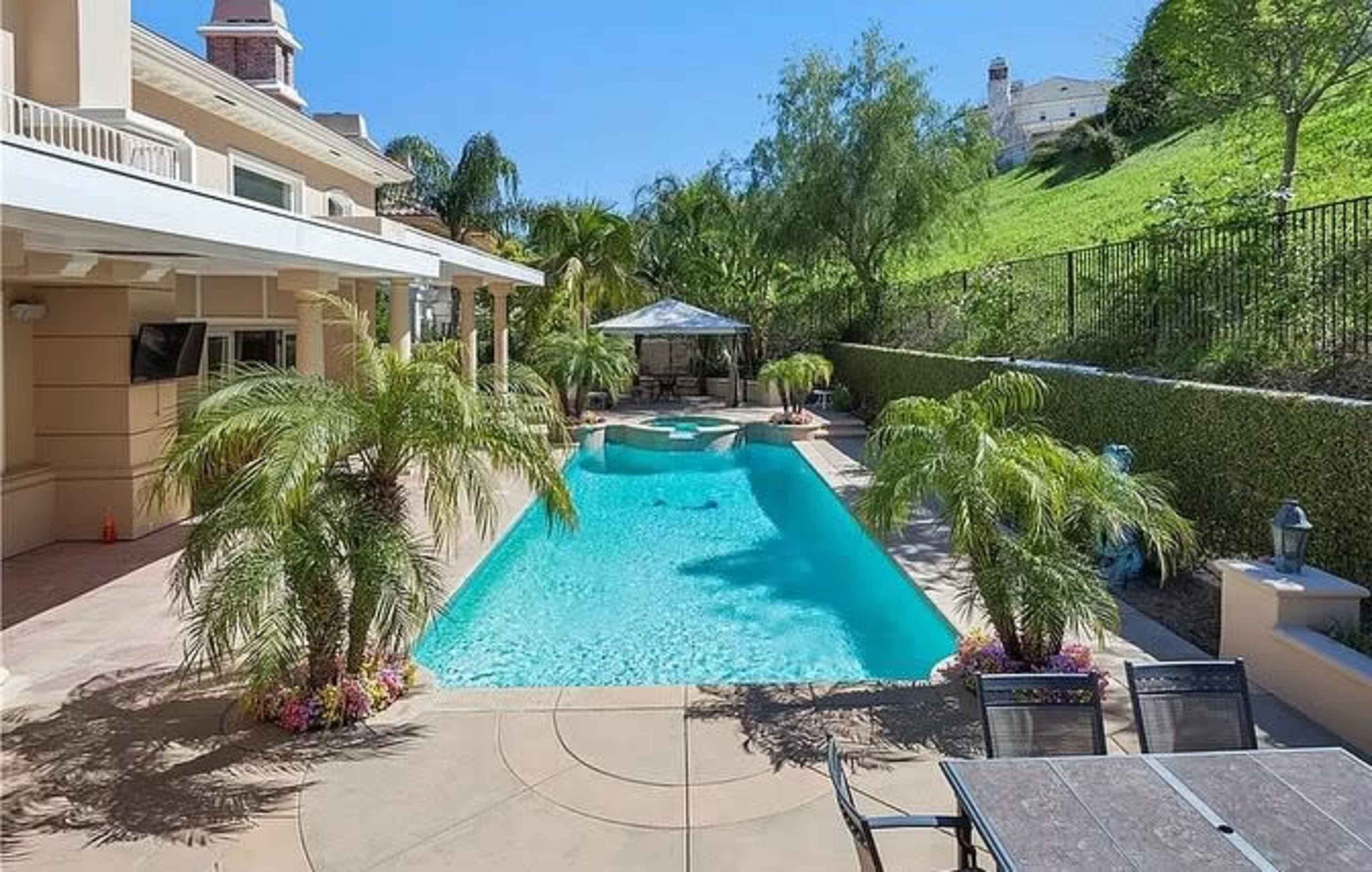 The image shows a residential backyard featuring a swimming pool surrounded by palm trees and a gazebo, with a hillside and fence in the background.