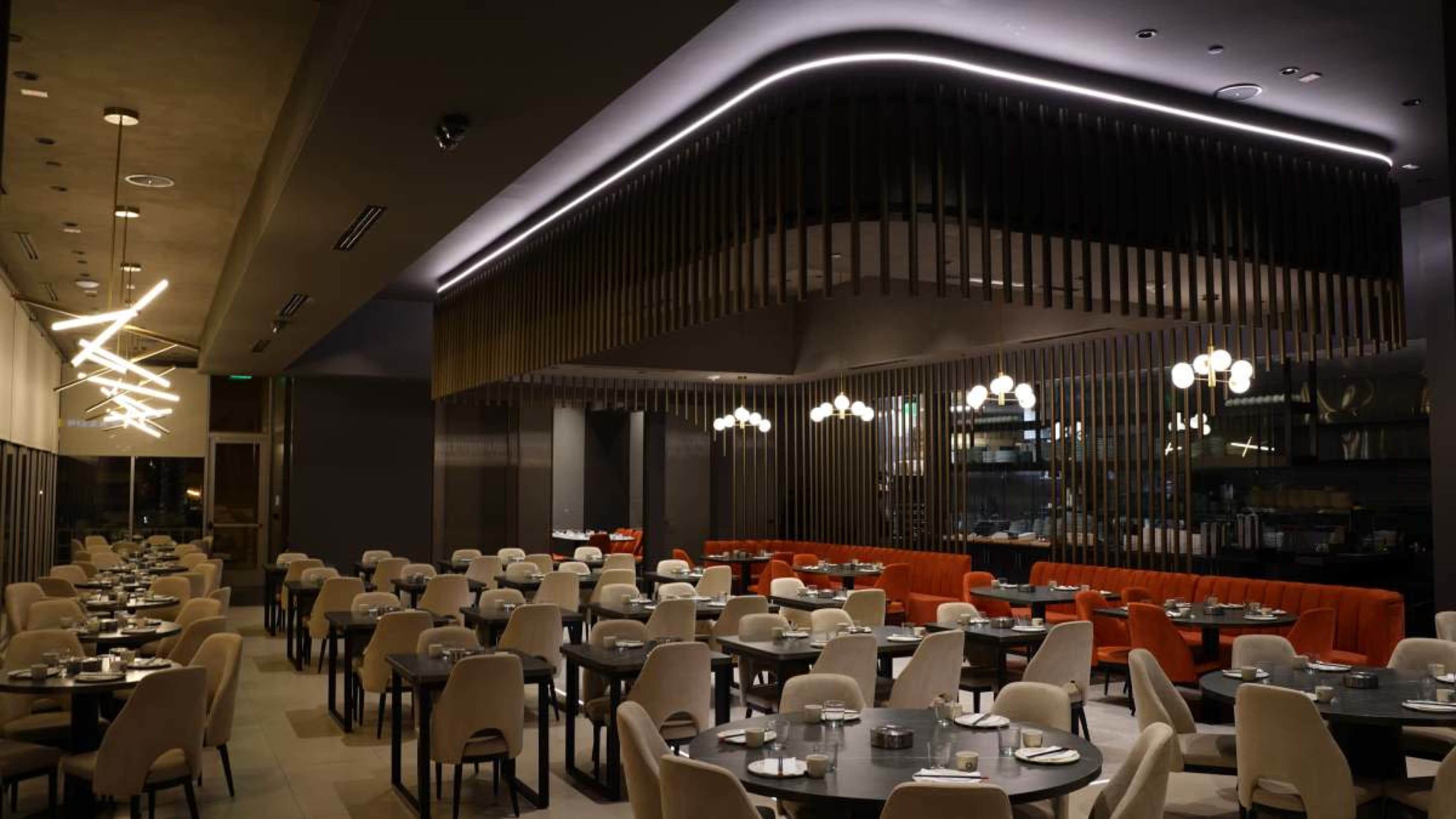 The image shows a modern restaurant interior with round tables set for dining, distinct lighting fixtures, and a combination of orange and neutral-colored seating.