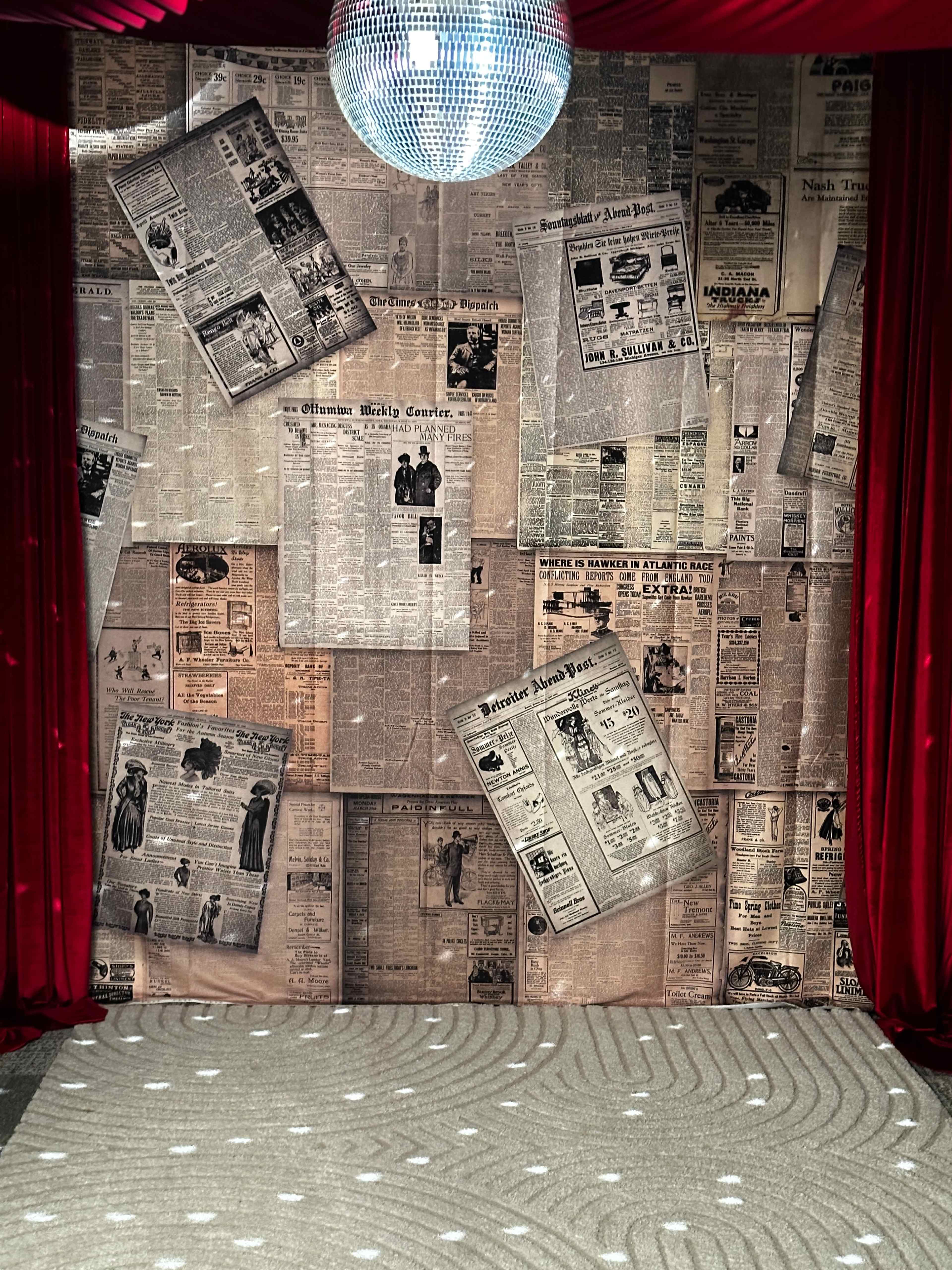 The image shows a backdrop covered in newspapers, with a disco ball hanging above and surrounded by red curtains.