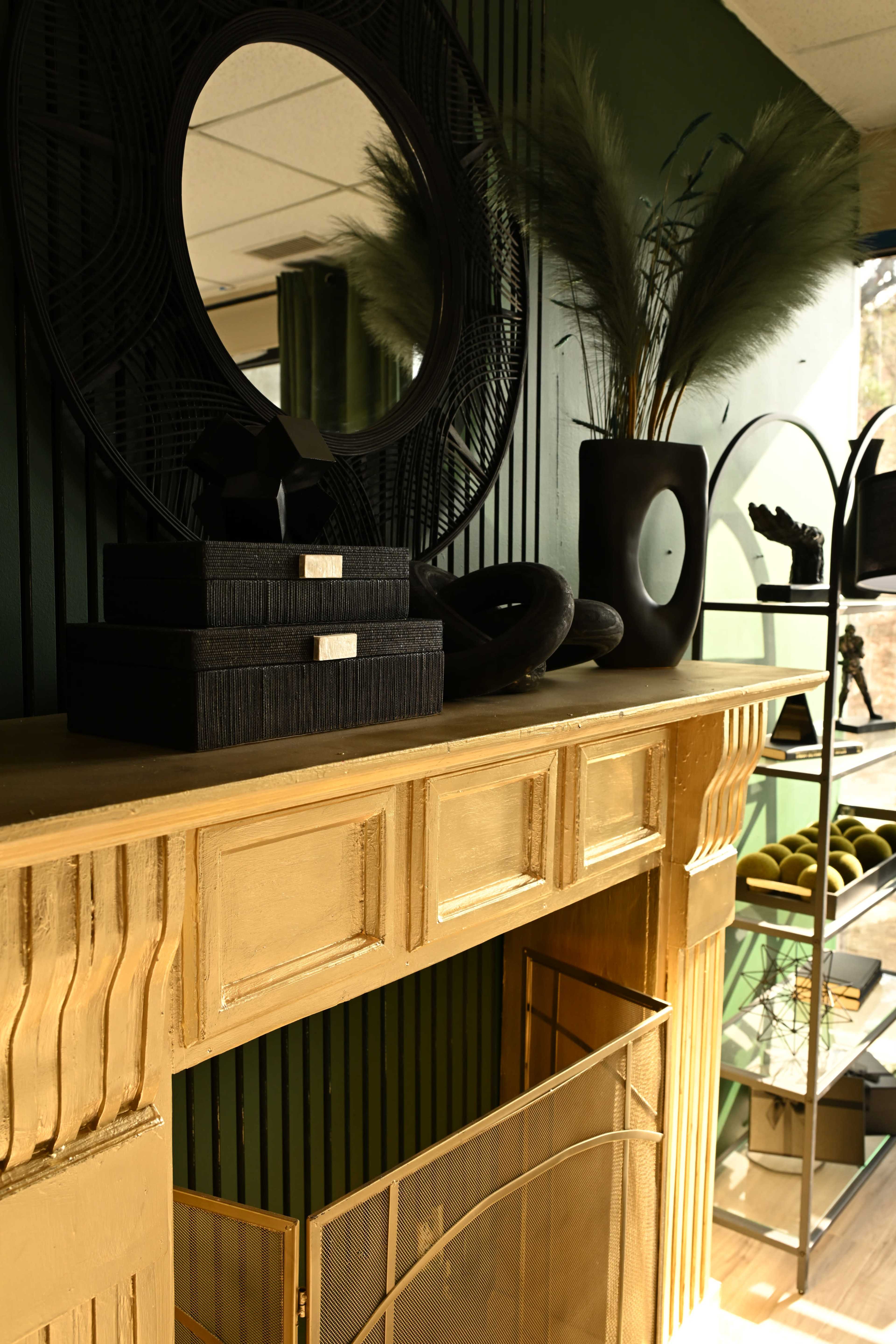 The image shows a stylized mantel with a gold finish, decorated with black boxes, a round mirror, and various decorative items against a dark green wall.