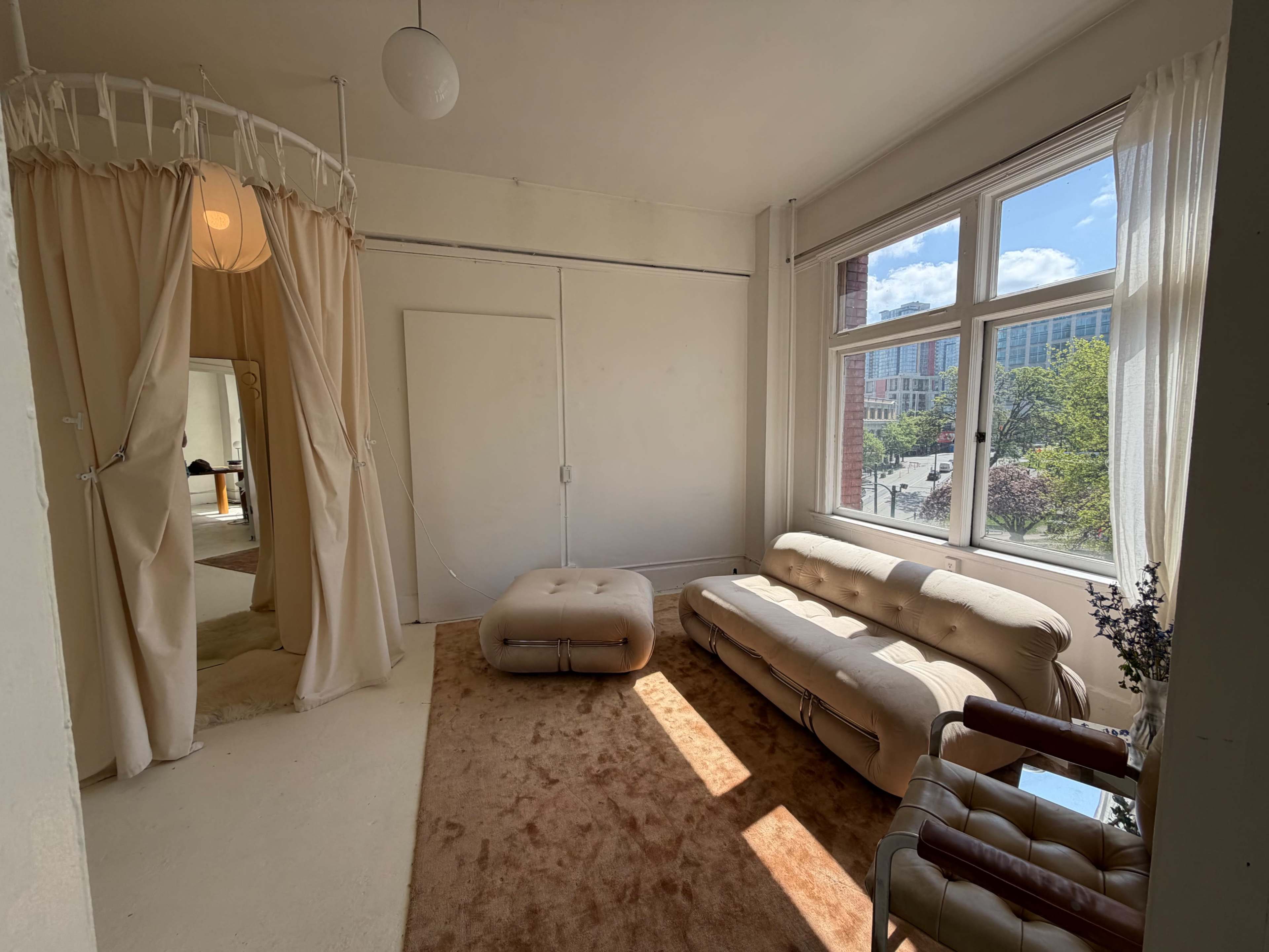 The image shows a bright, minimalist room with a beige couch, a large window letting in natural light, and draped curtains creating a partitioned area.