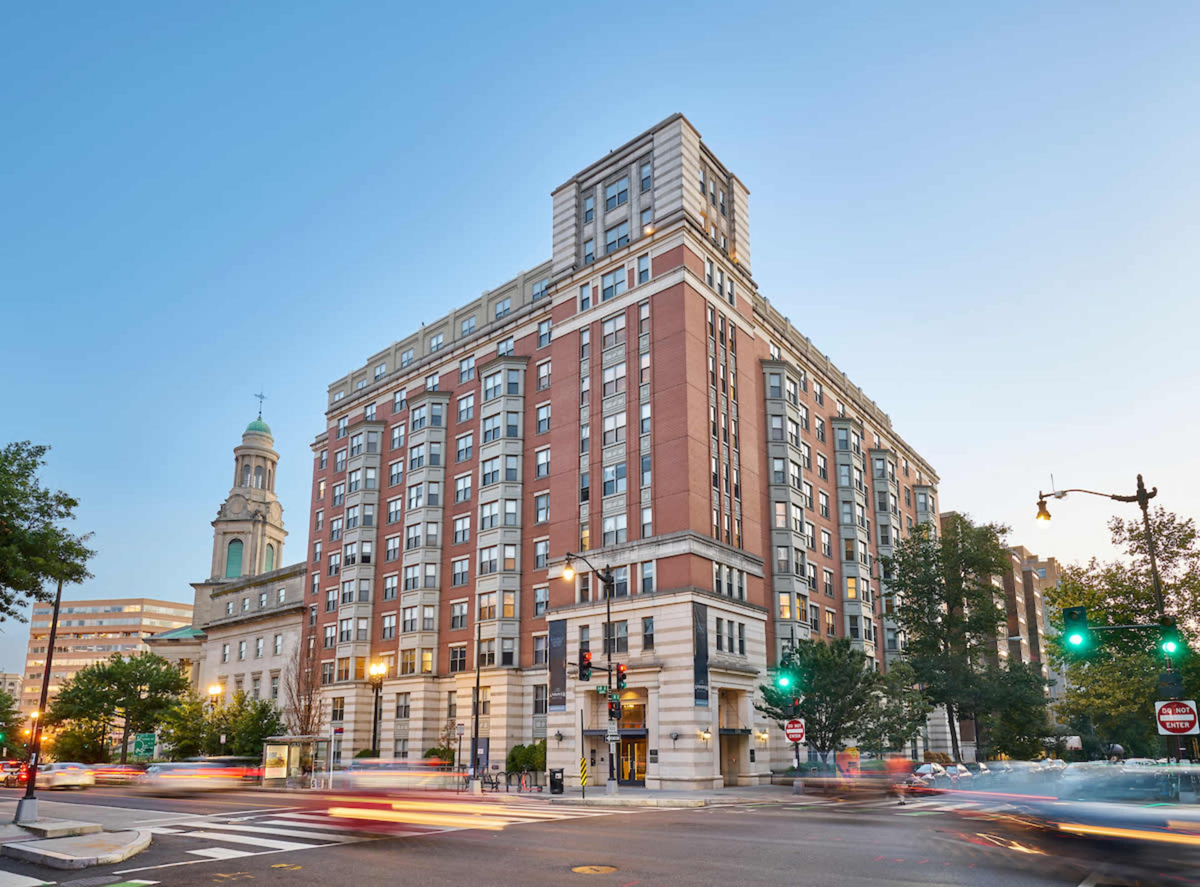 The image shows a multi-story brick building at dusk, with a clock tower visible in the background and traffic passing by.