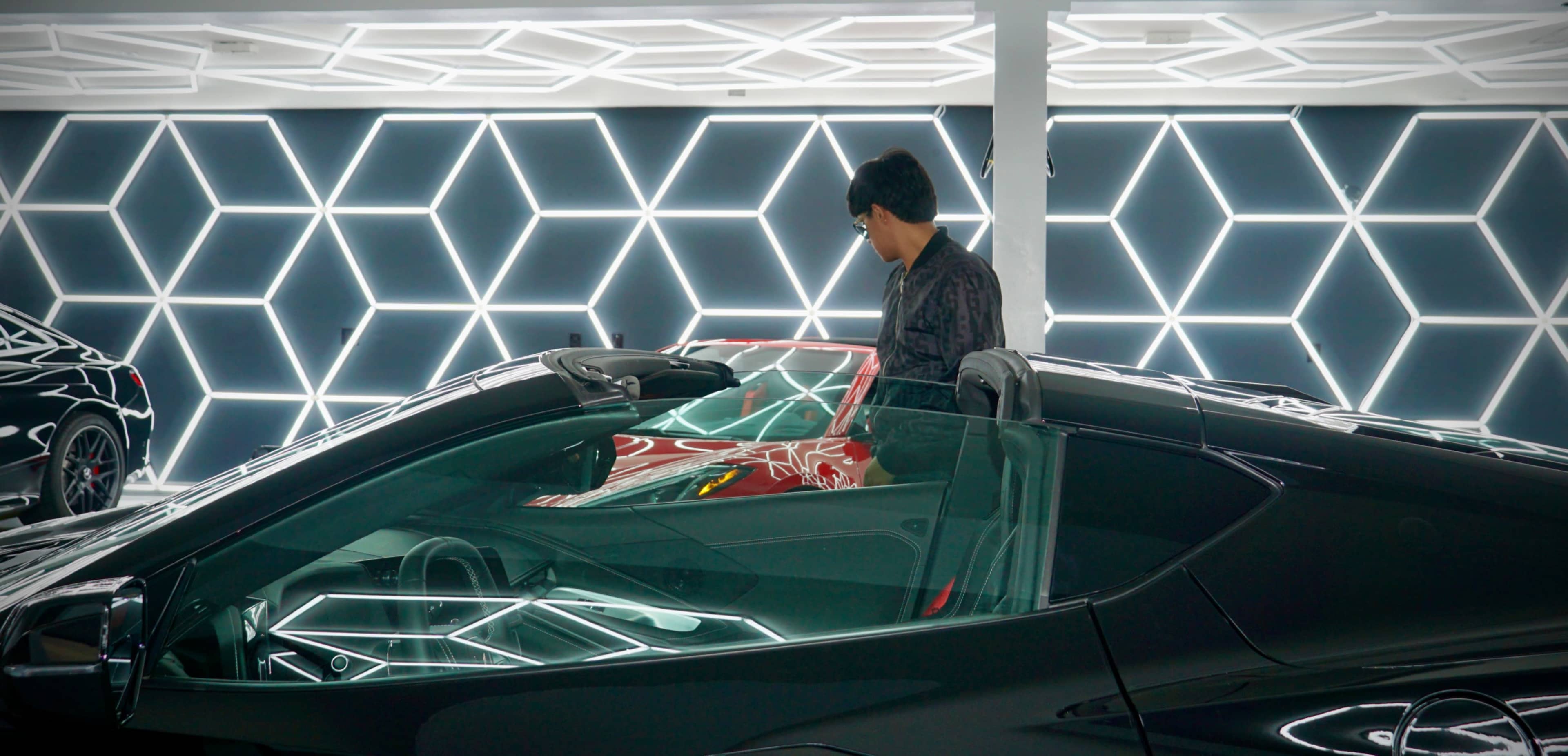 A person stands next to a luxury convertible car in a brightly illuminated, angular-patterned garage.