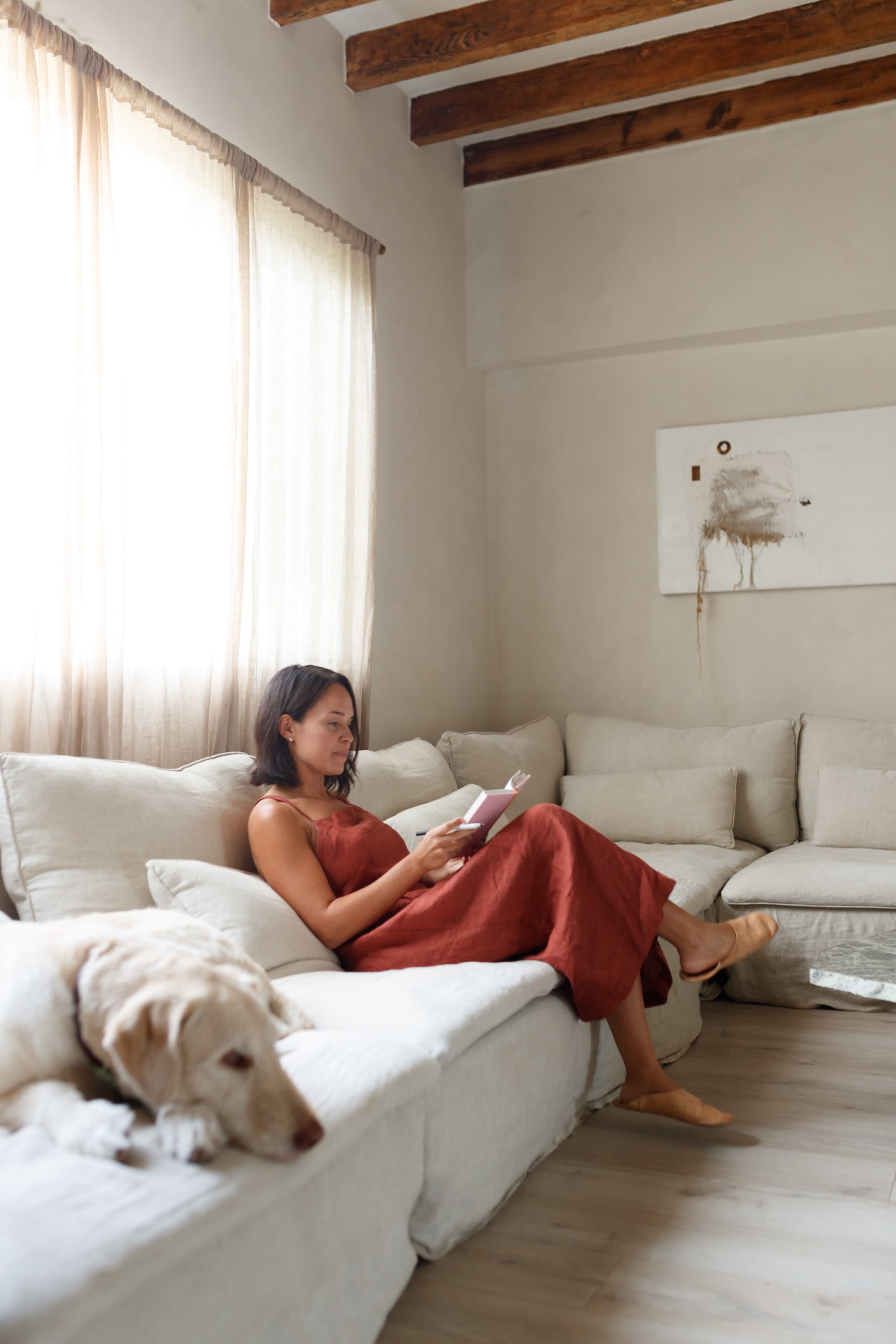 A woman in a red dress sits on a light-colored sofa, reading a book while a dog lies nearby.