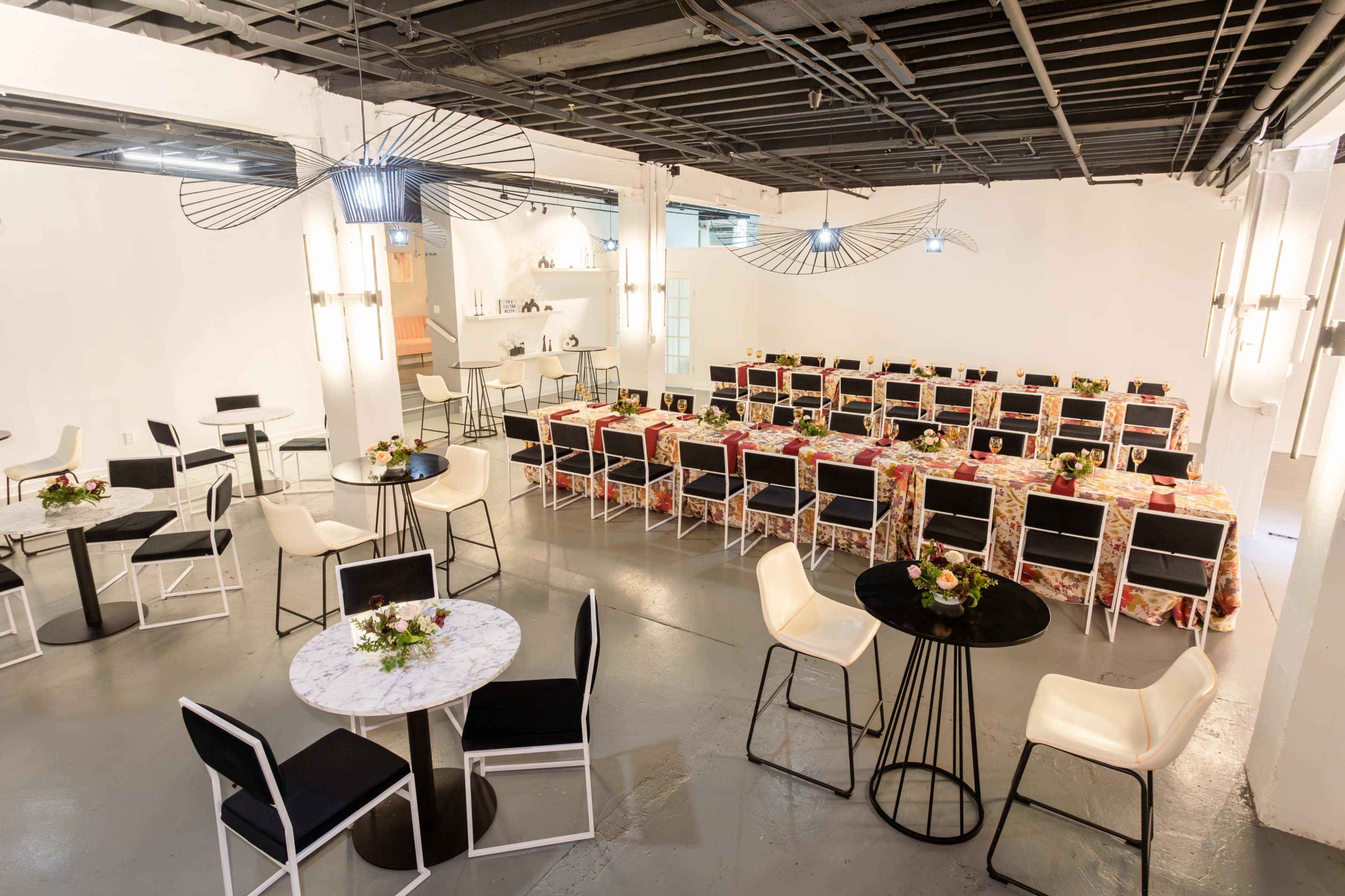 A spacious event room is set with rows of tables covered in patterned cloths, surrounded by black and white chairs, and decorated with small floral centerpieces.