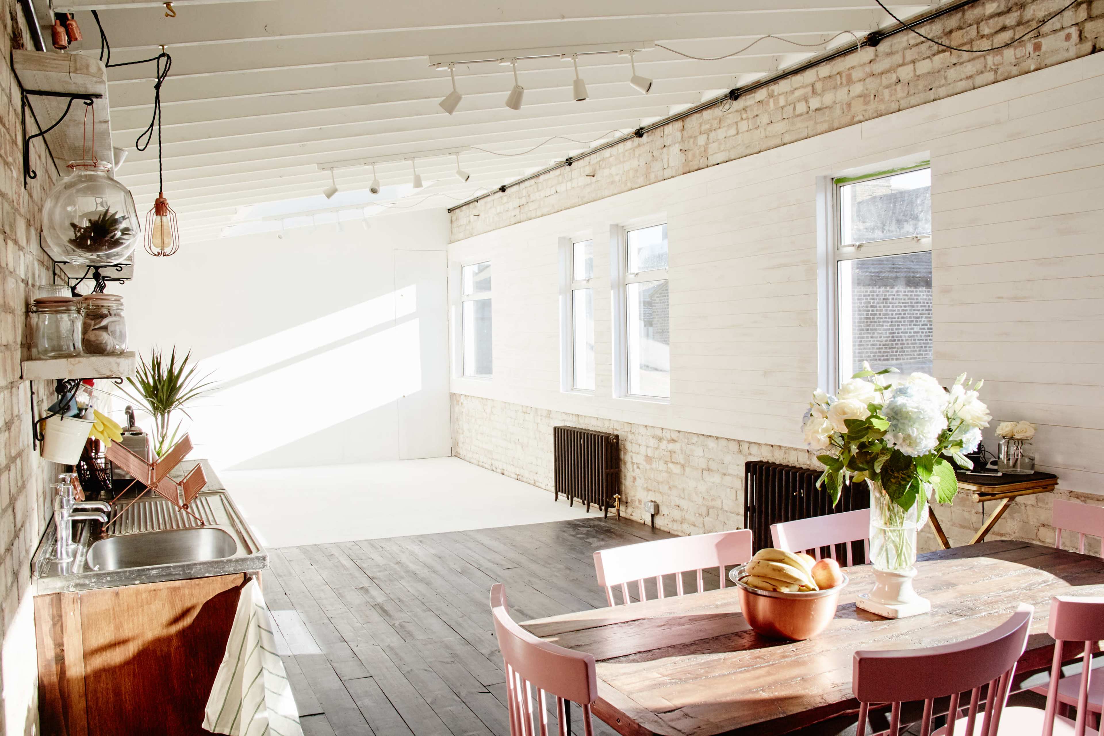 A bright, airy room features a cozy kitchen area with a sink and a table set with a vase of flowers and fruit, alongside large windows allowing natural light to fill the space.