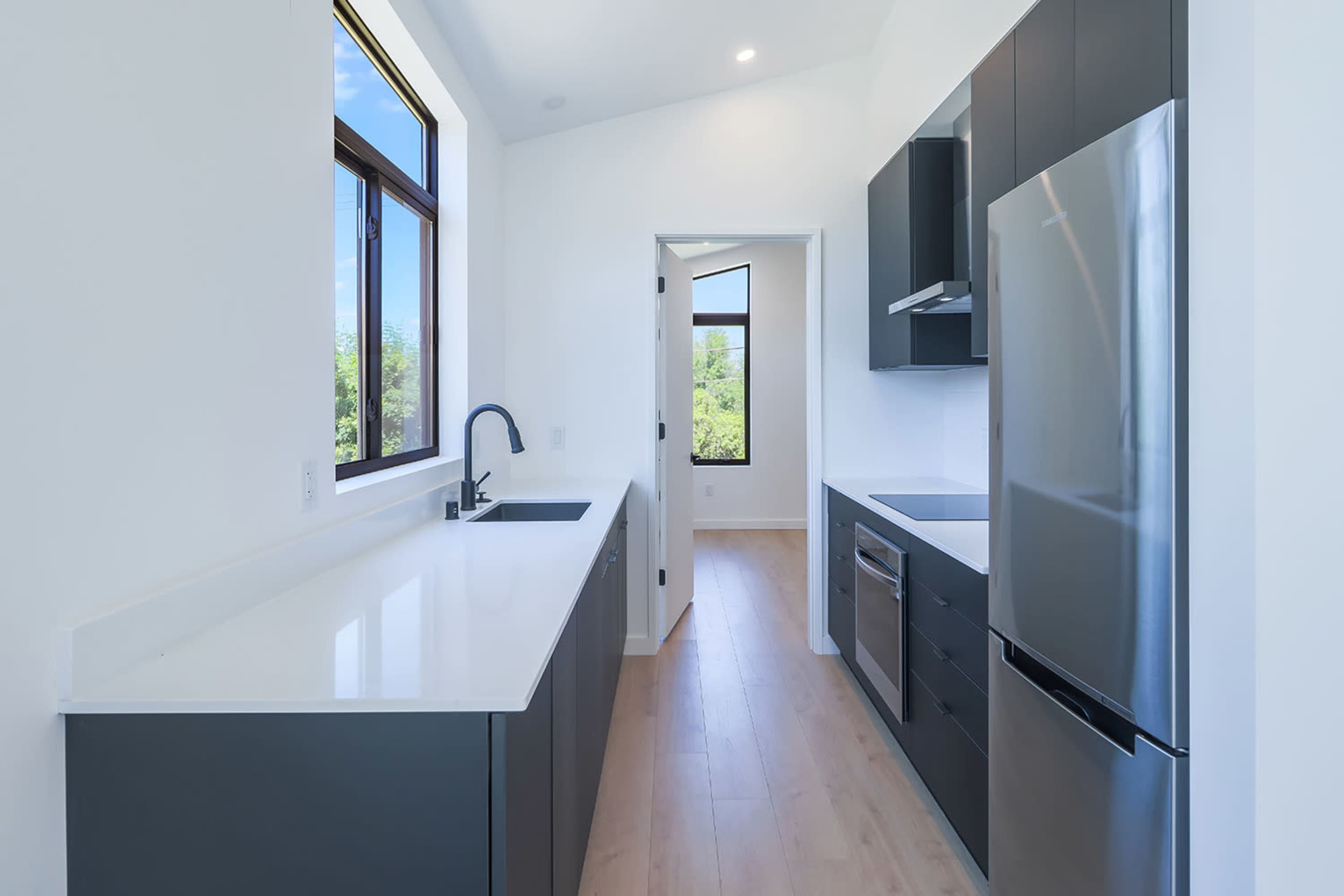 A narrow kitchen features sleek cabinetry, a stainless steel refrigerator, and a large window allowing natural light.