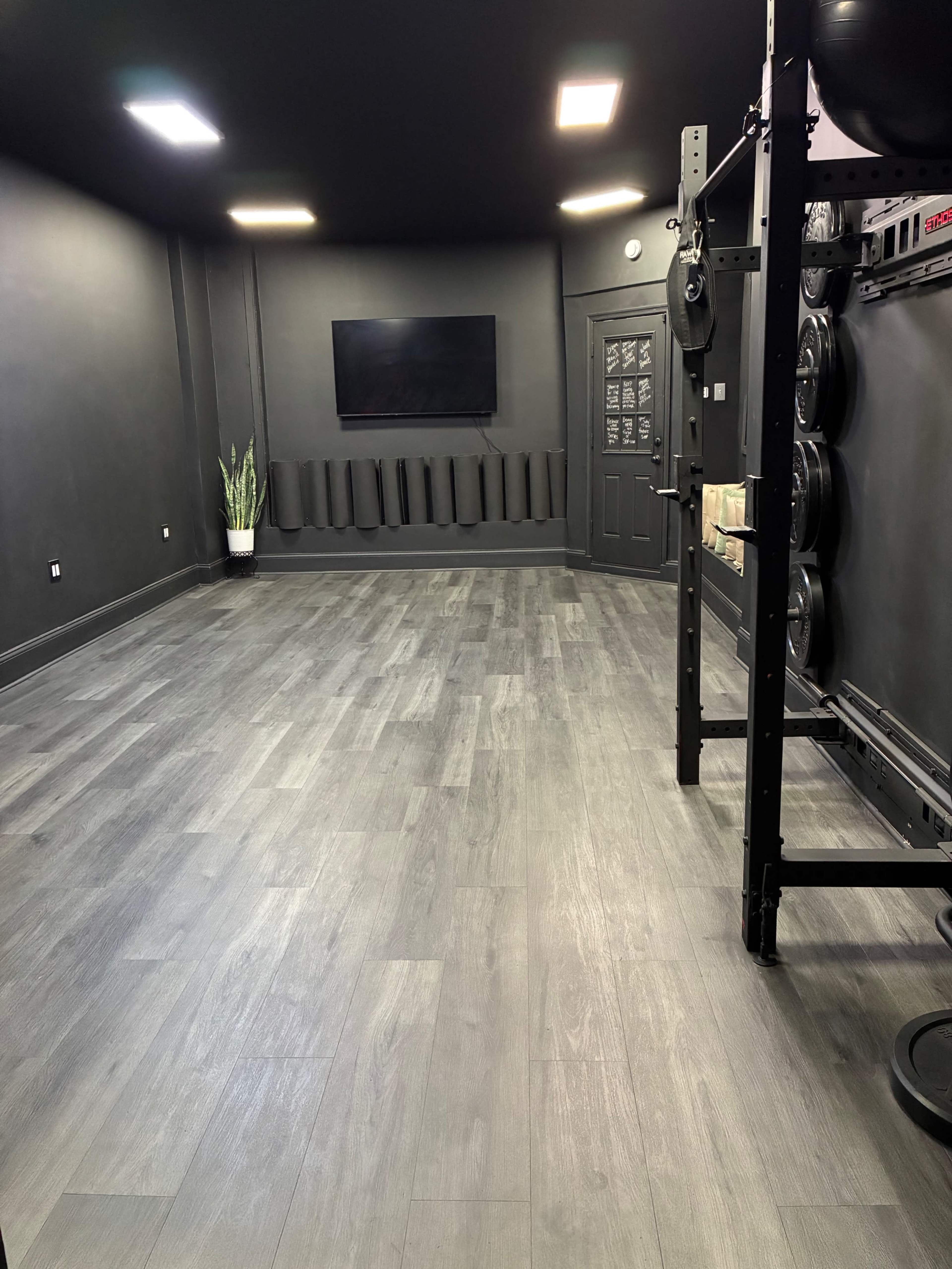 The image features a minimalist, dark-colored gym space with wooden flooring, a wall-mounted television, and a weight rack on the right side.