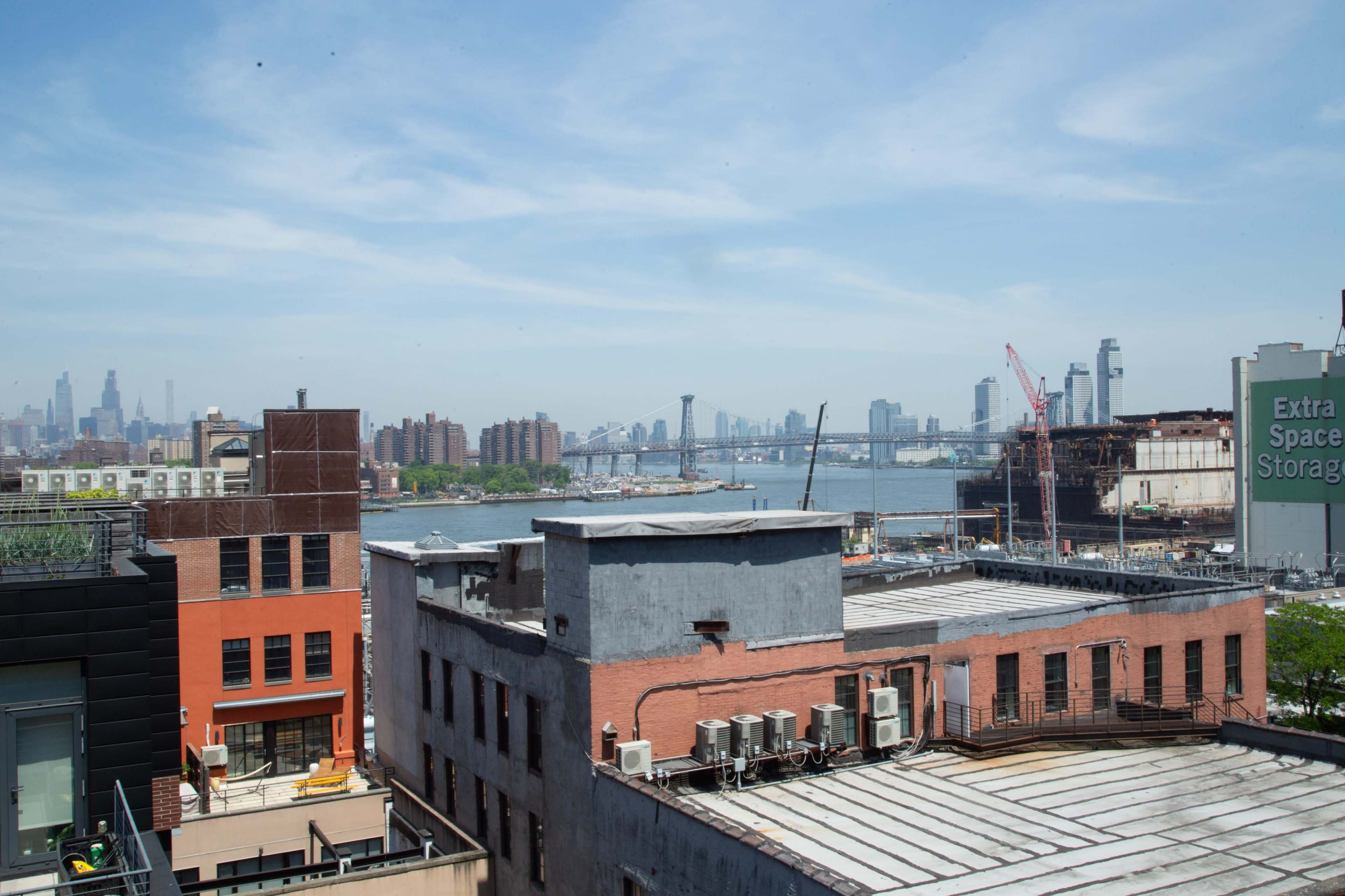 The image shows a view of buildings and rooftops in the foreground, with a river and a bridge in the background against a clear sky.
