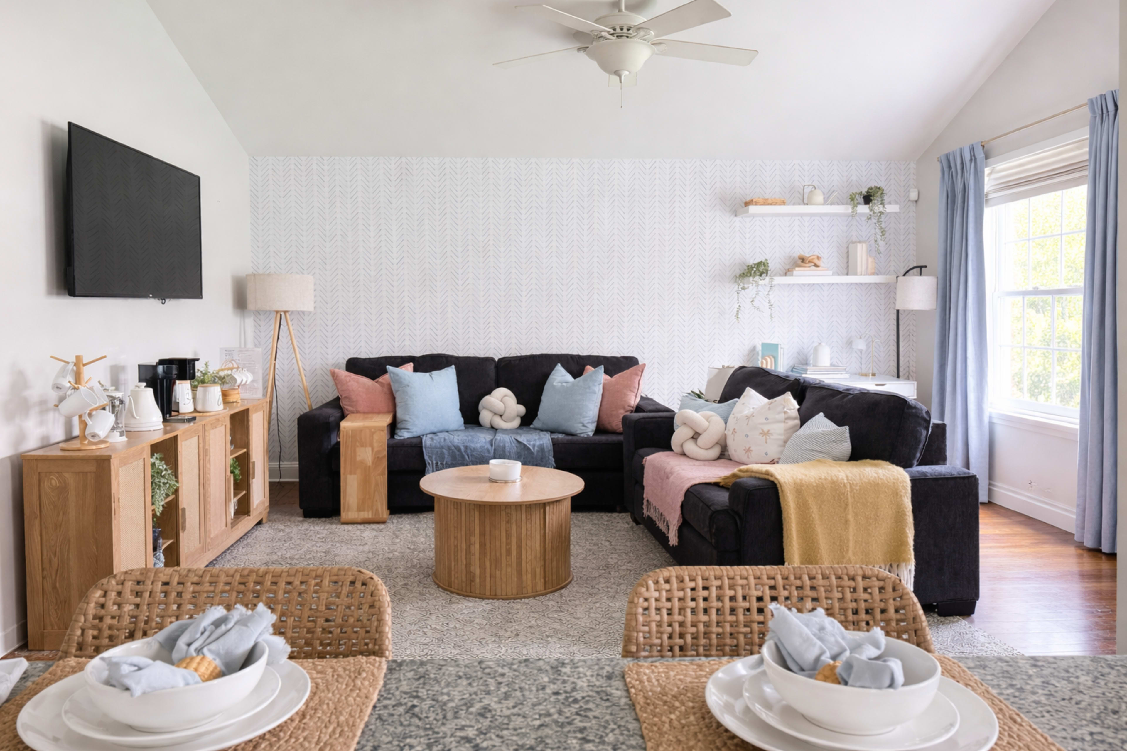 The image shows a modern living room with two sofas, a round coffee table, a television mounted on the wall, and decorative shelves.