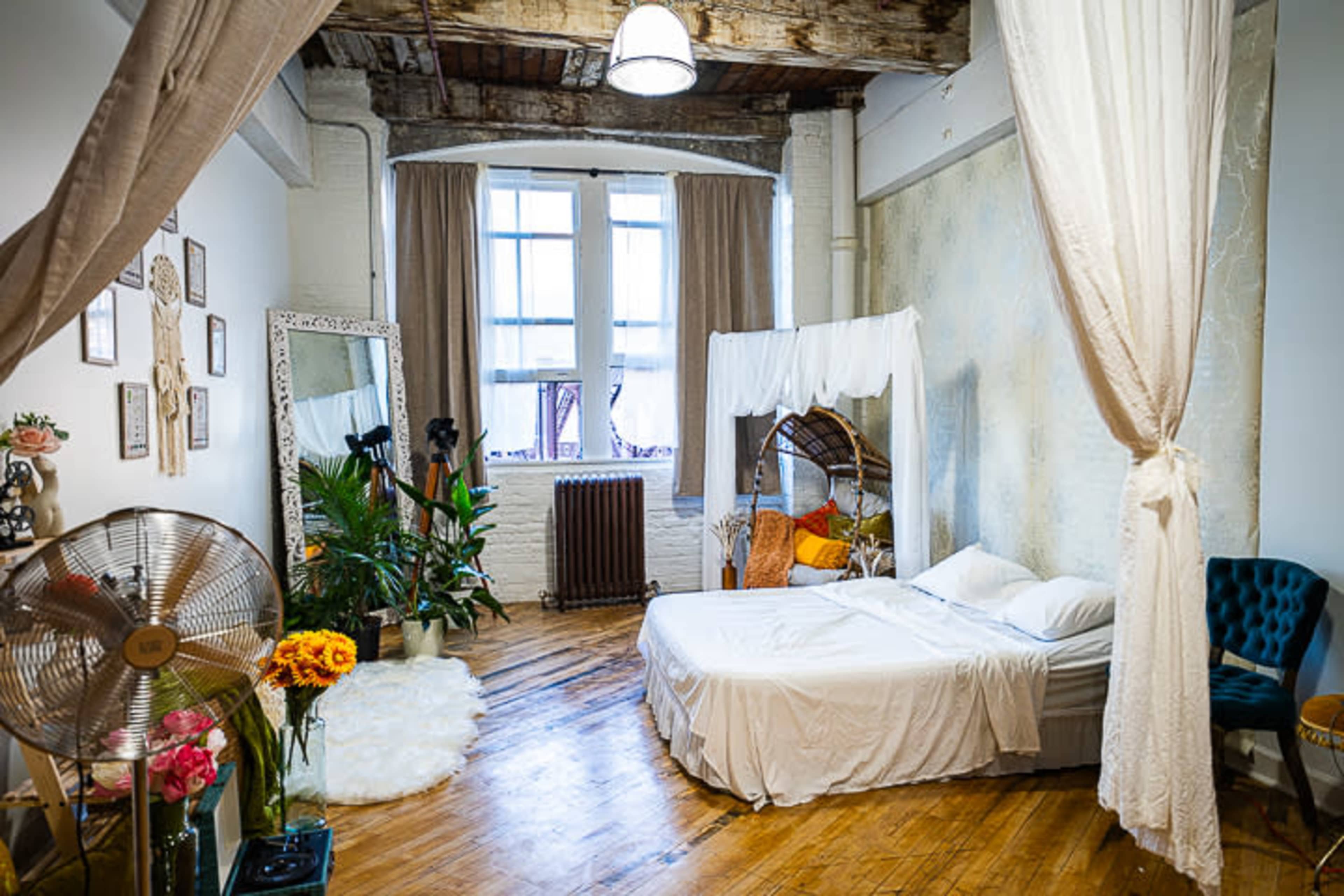 A bright bedroom featuring a bed with white linens, a mirror, plants, and a rustic wooden floor.