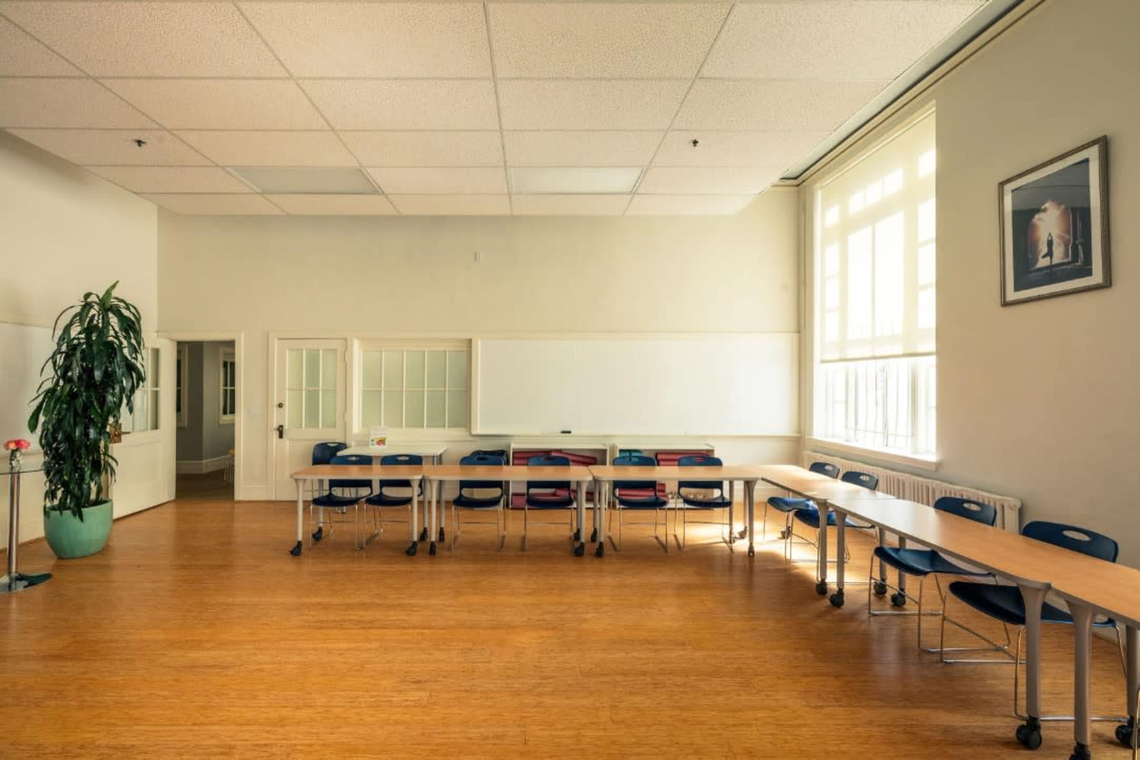 A sparsely furnished classroom with a long table and several chairs arranged around it, along with a whiteboard and a large window providing natural light.