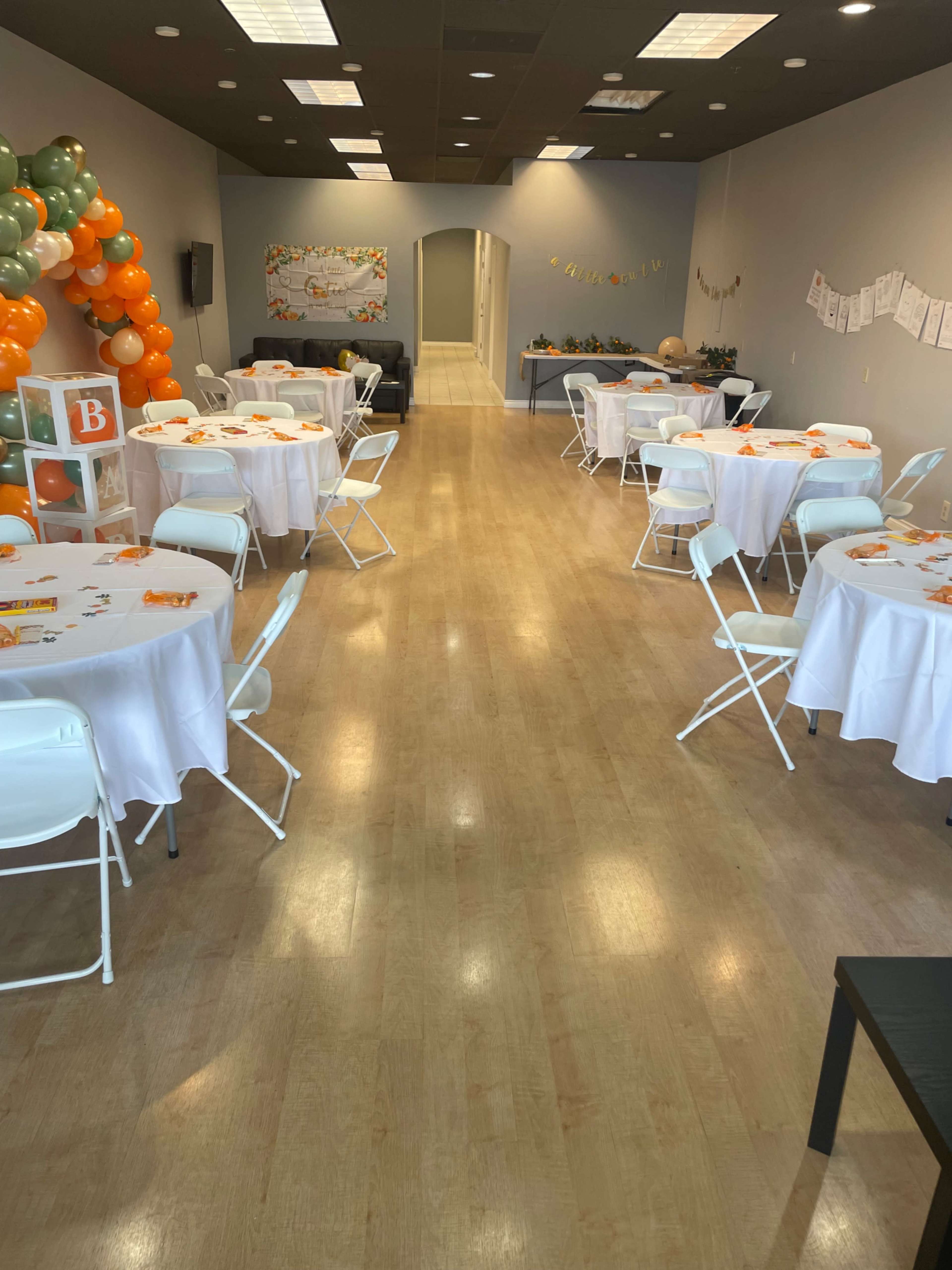 The image shows a spacious event room set up with several decorated tables and an arch of orange and green balloons.