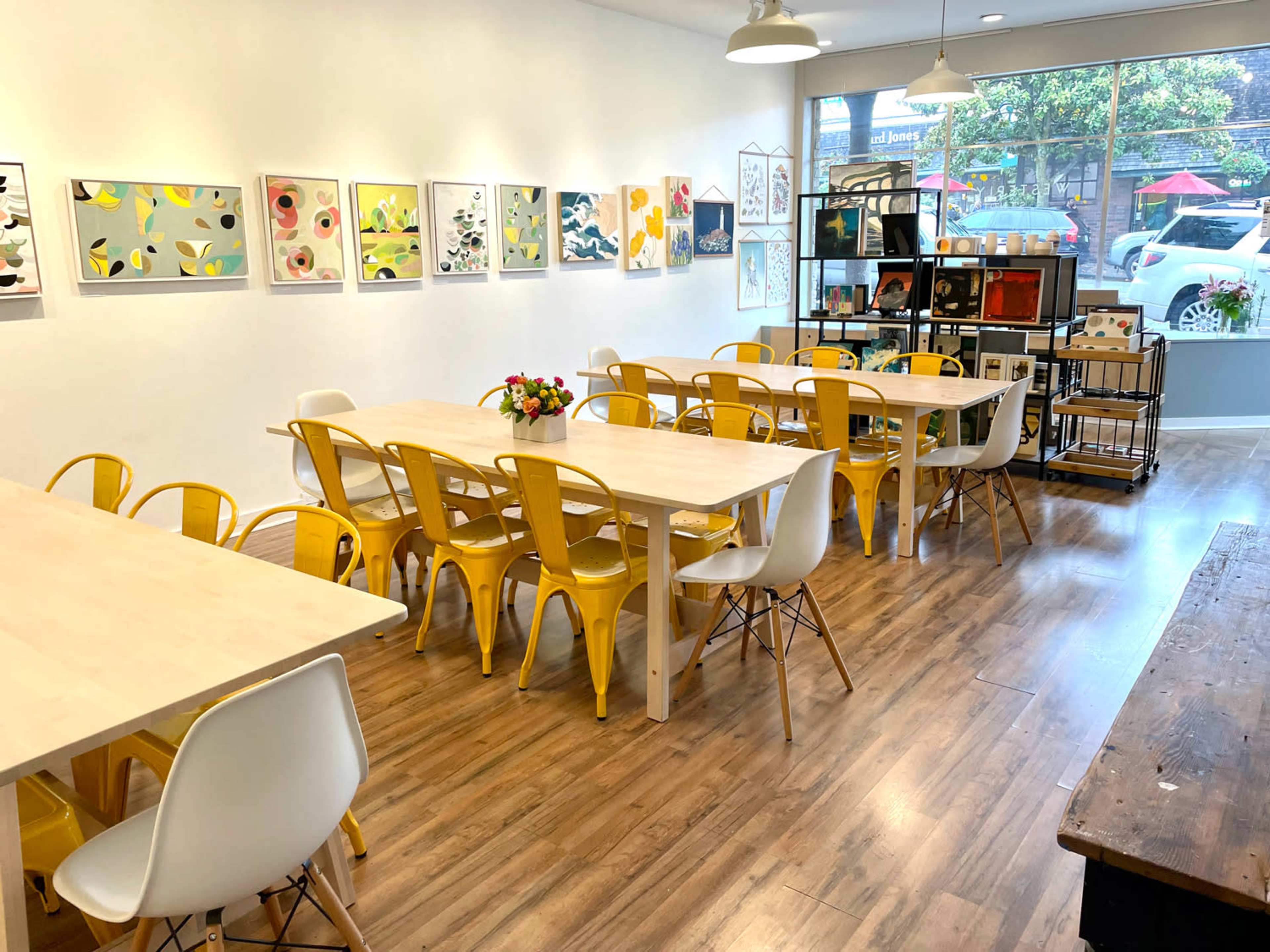 A brightly lit room featuring multiple tables and chairs, with art displayed on the walls and a shelving unit filled with various items.