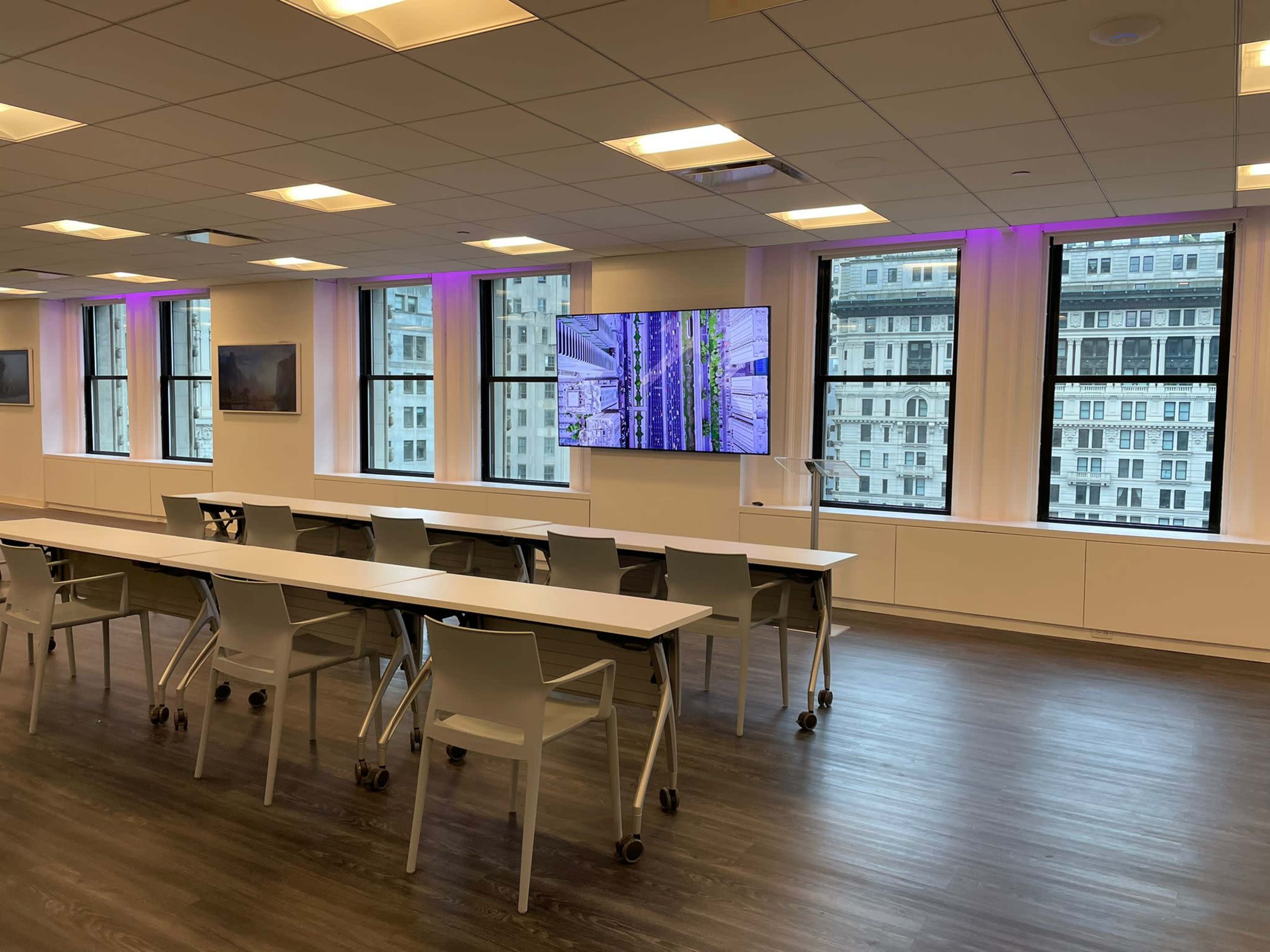 A modern conference room features a long table with rolling chairs and a large screen displaying visuals, illuminated by purple lighting from the windows.