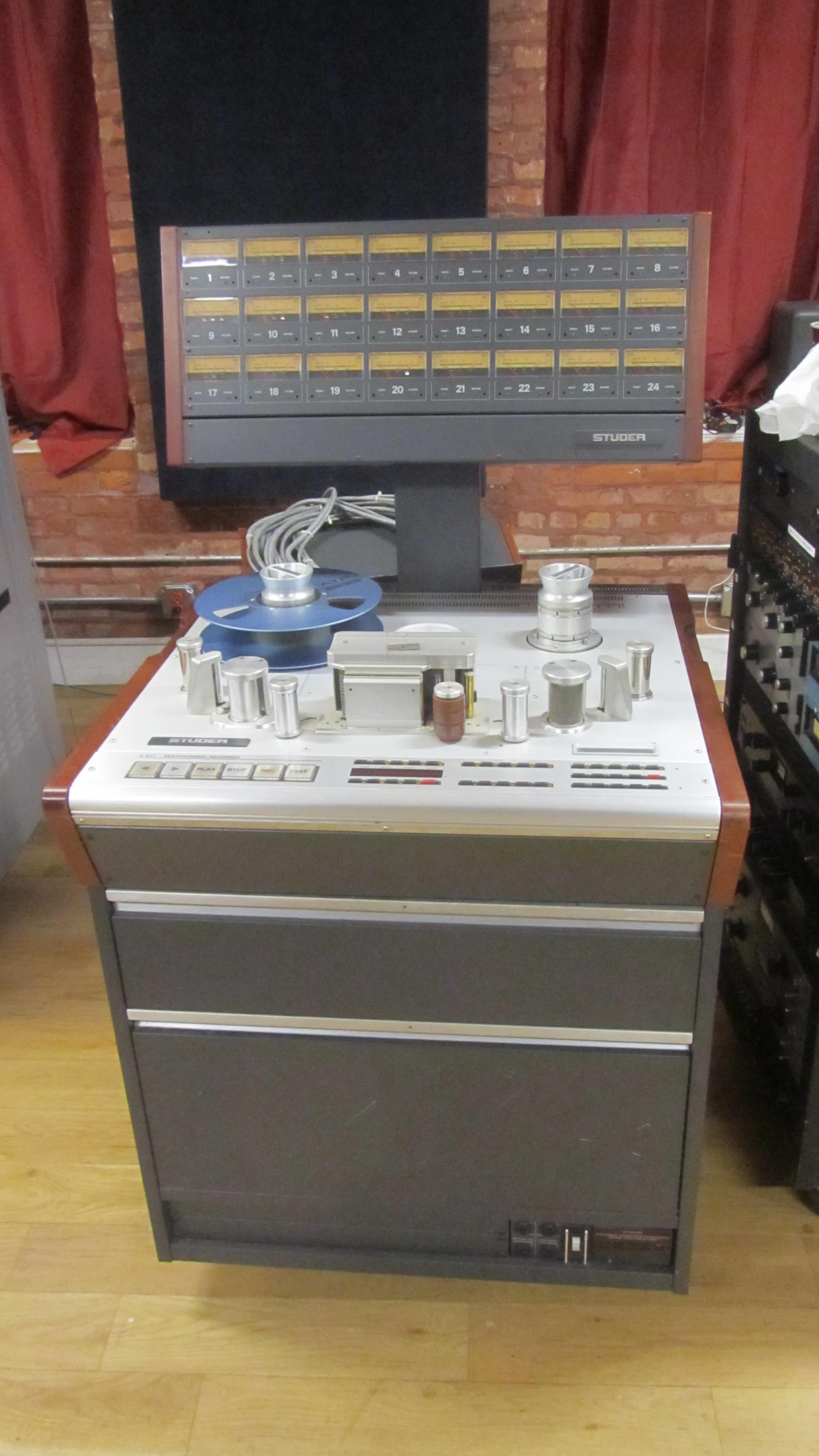 A vintage audio recording console with a large control panel and reels for tape playback sits on a wooden floor against a brick wall.