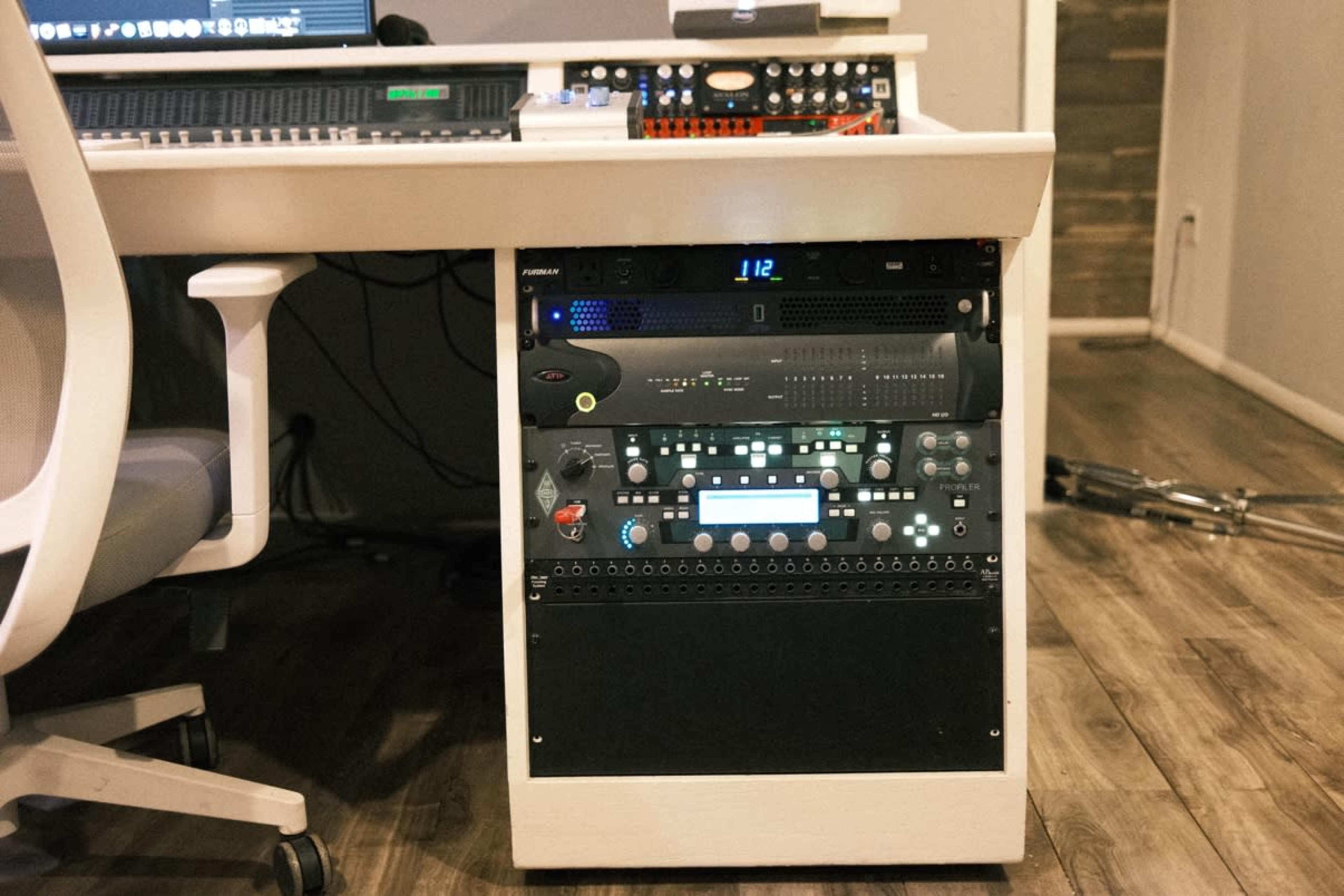 The image shows a mounted audio equipment rack with various electronic devices and controls, positioned beside a workstation in a studio setting.