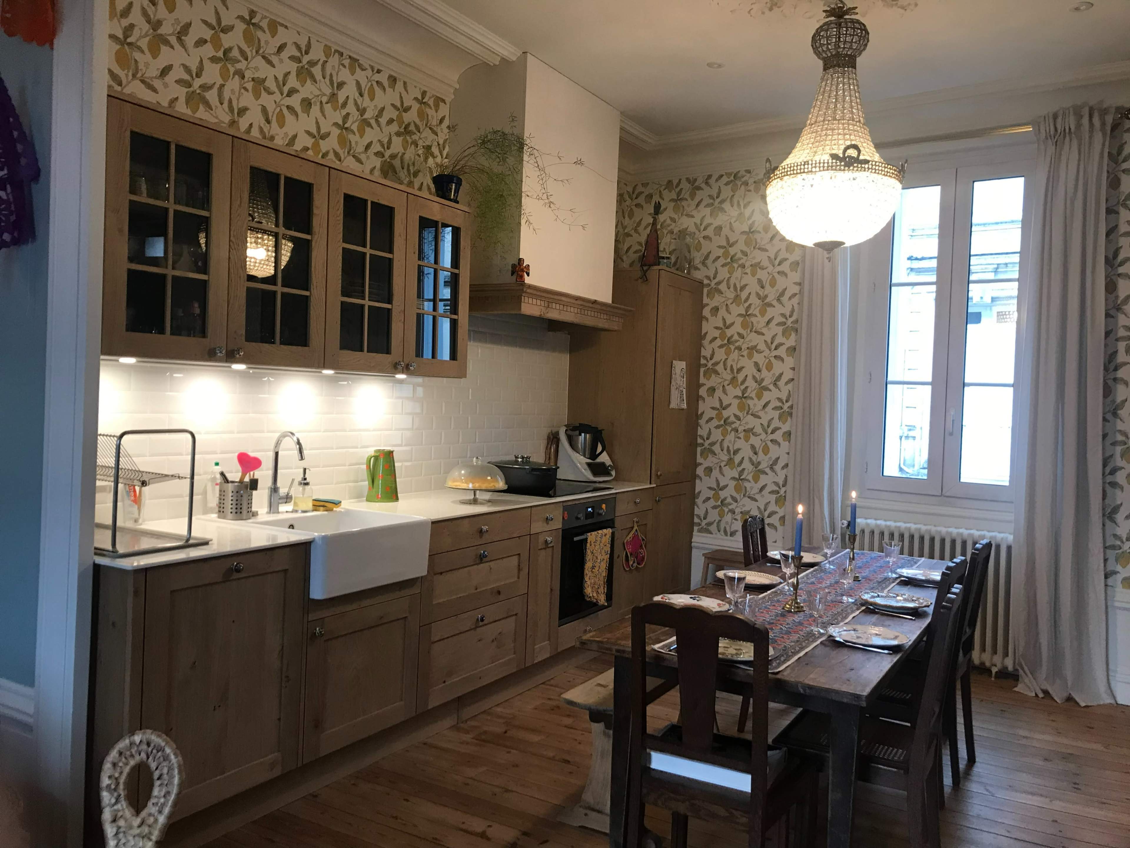 The image shows a kitchen and dining area featuring wooden cabinetry, a large farmhouse sink, and a long wooden dining table set for several people, with floral wallpaper and a chandelier overhead.