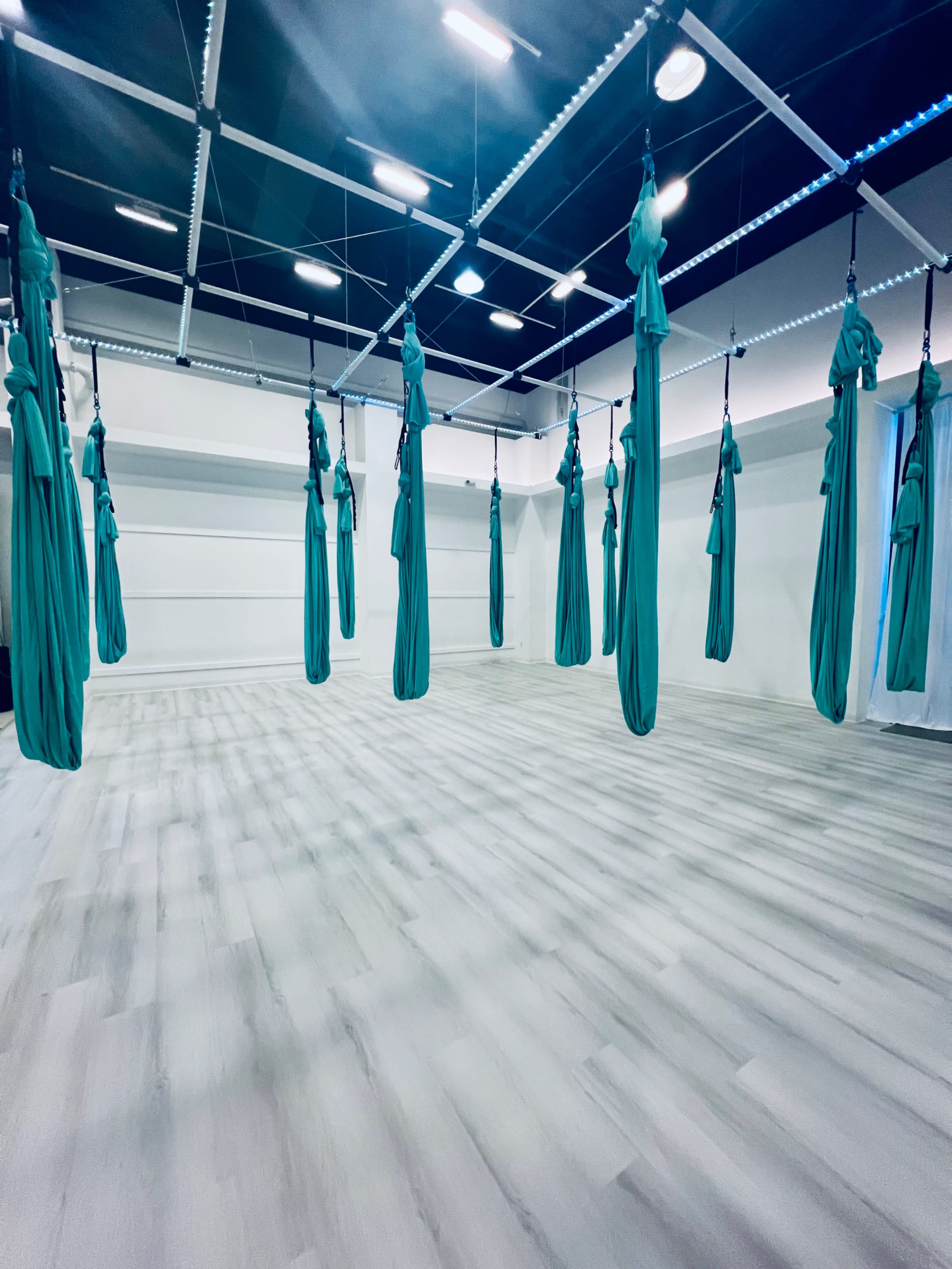 The image shows an indoor studio with several teal aerial silks hanging from the ceiling, arranged in a spacious, brightly lit room.