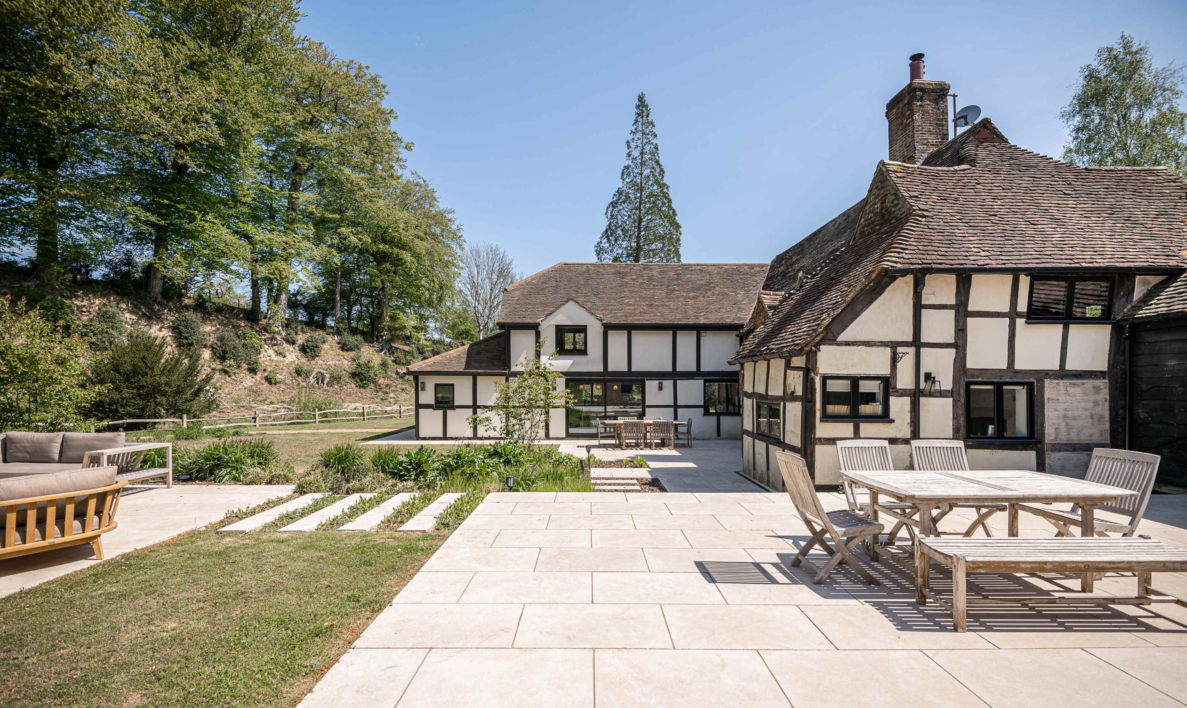 A traditional half-timbered house is situated beside a modern extension, with a stone patio and outdoor seating area surrounded by greenery.