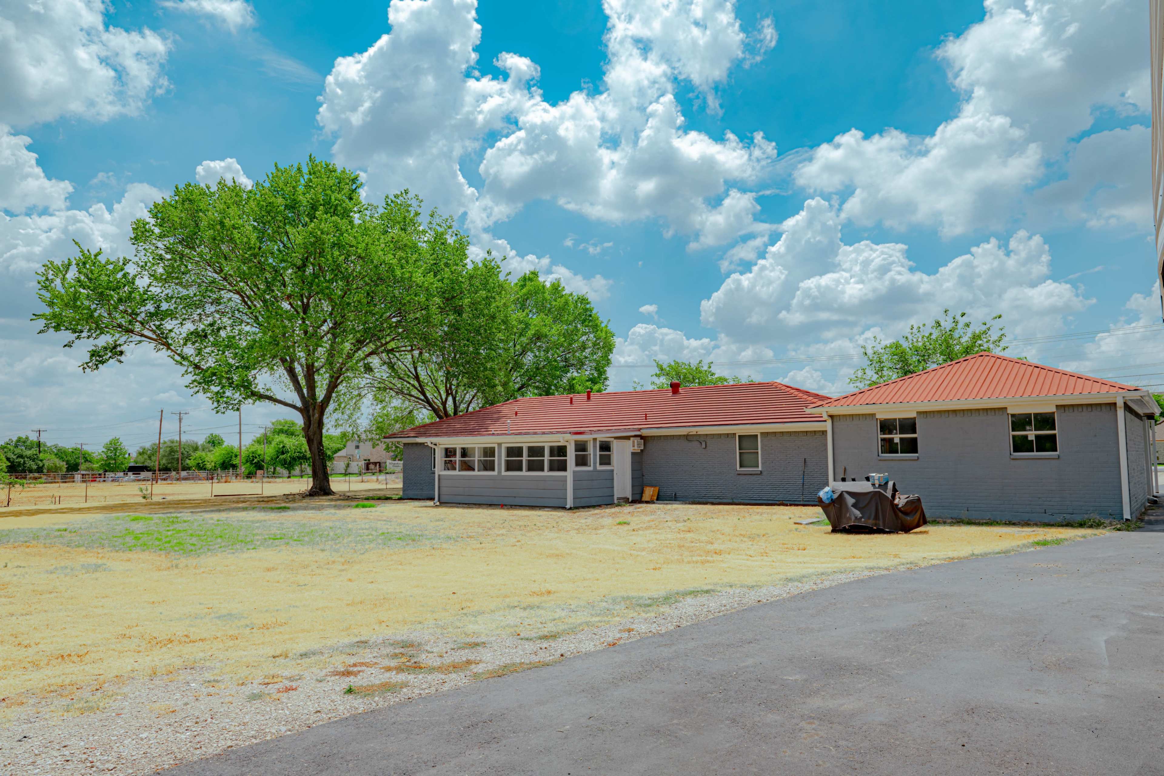 A gray house with a red metal roof is situated next to a large tree in an open, grassy area under a cloudy blue sky.