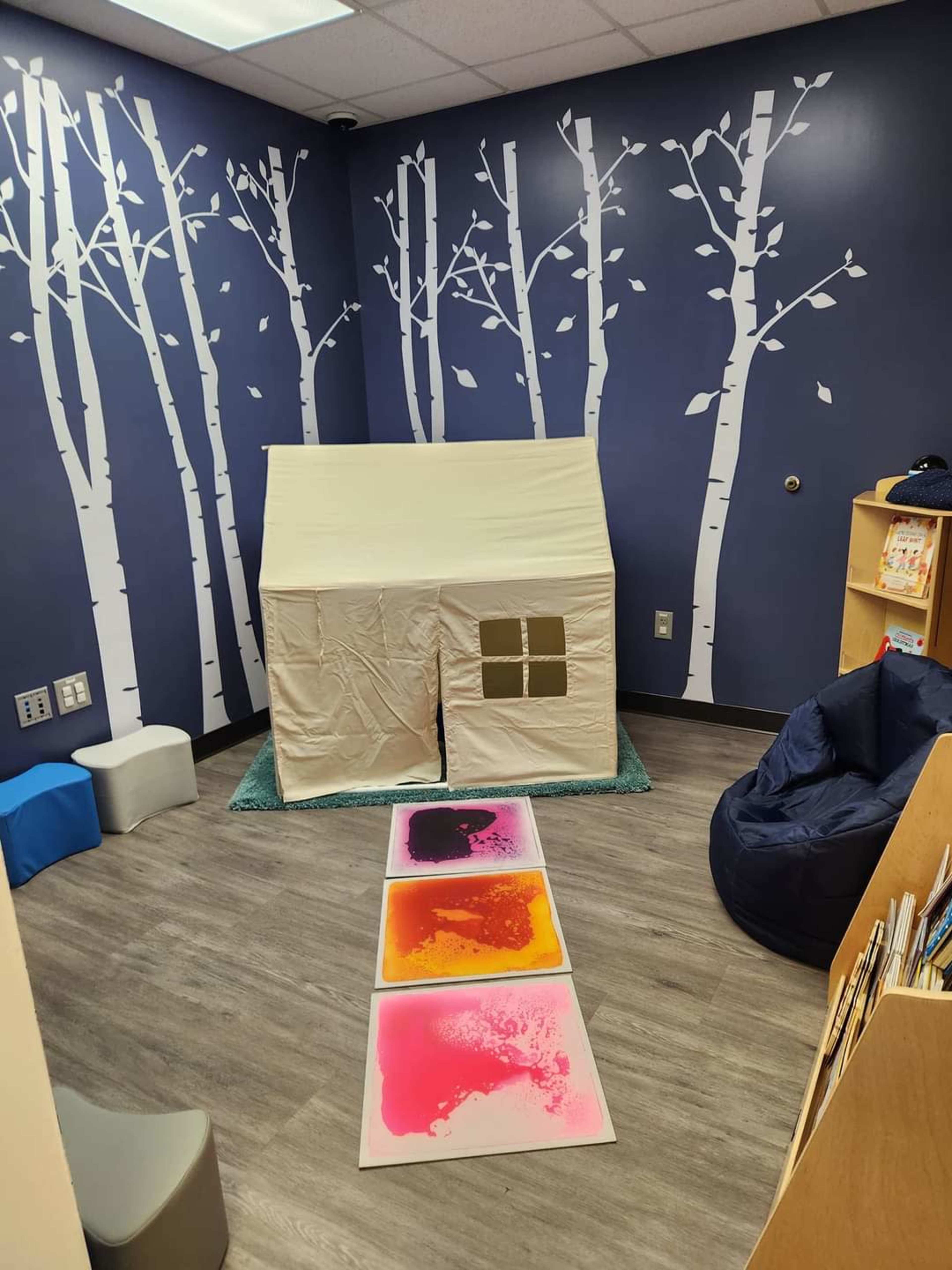 A small playroom featuring a tent-like structure, colorful mats, and a blue bean bag chair against a wall decorated with white trees.