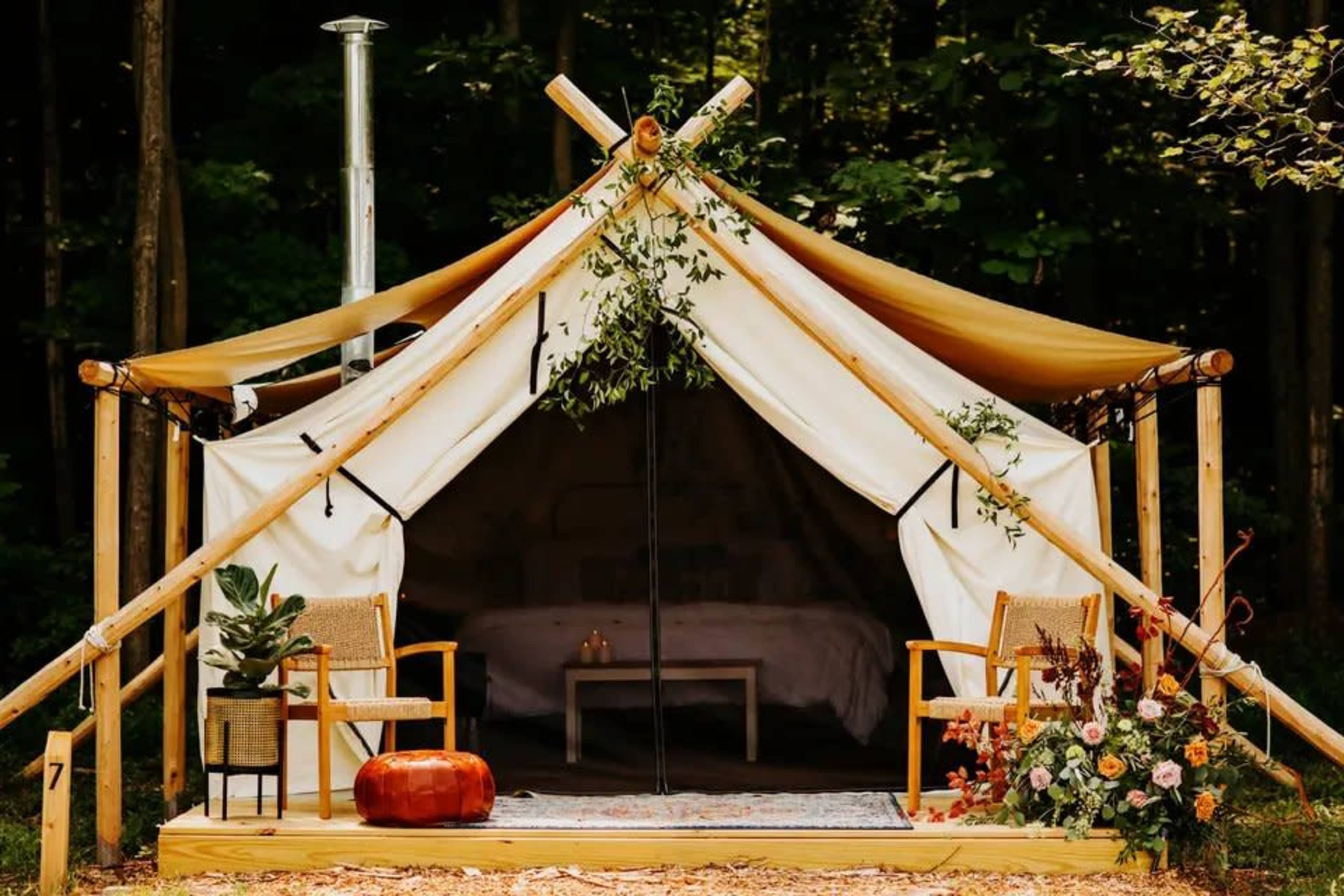 A glamping tent with a wooden porch, two chairs, and decorative plants is set amid a wooded area.