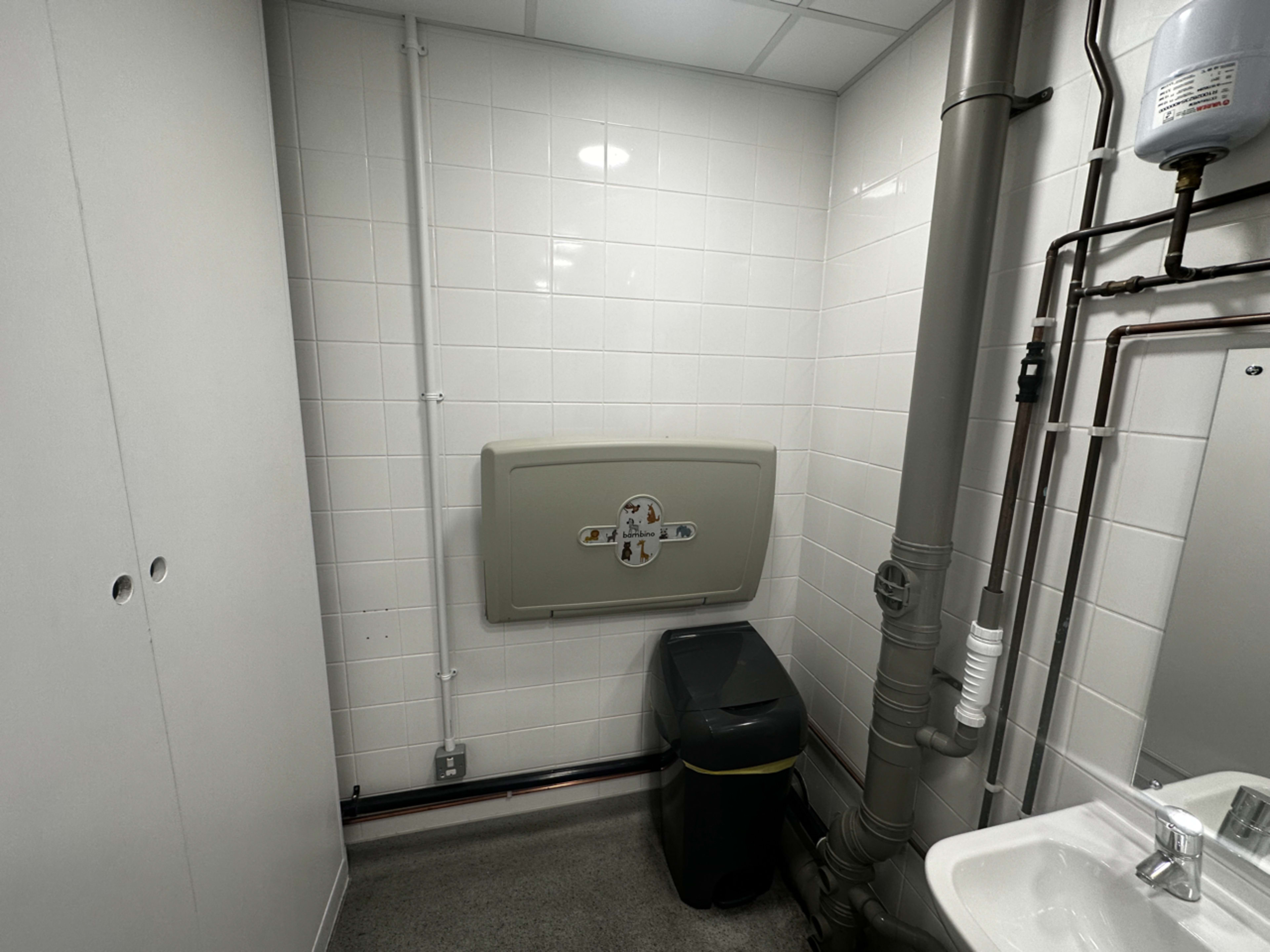The image shows a simple bathroom interior featuring a toilet, a sink, a trash bin, and exposed plumbing along the walls.
